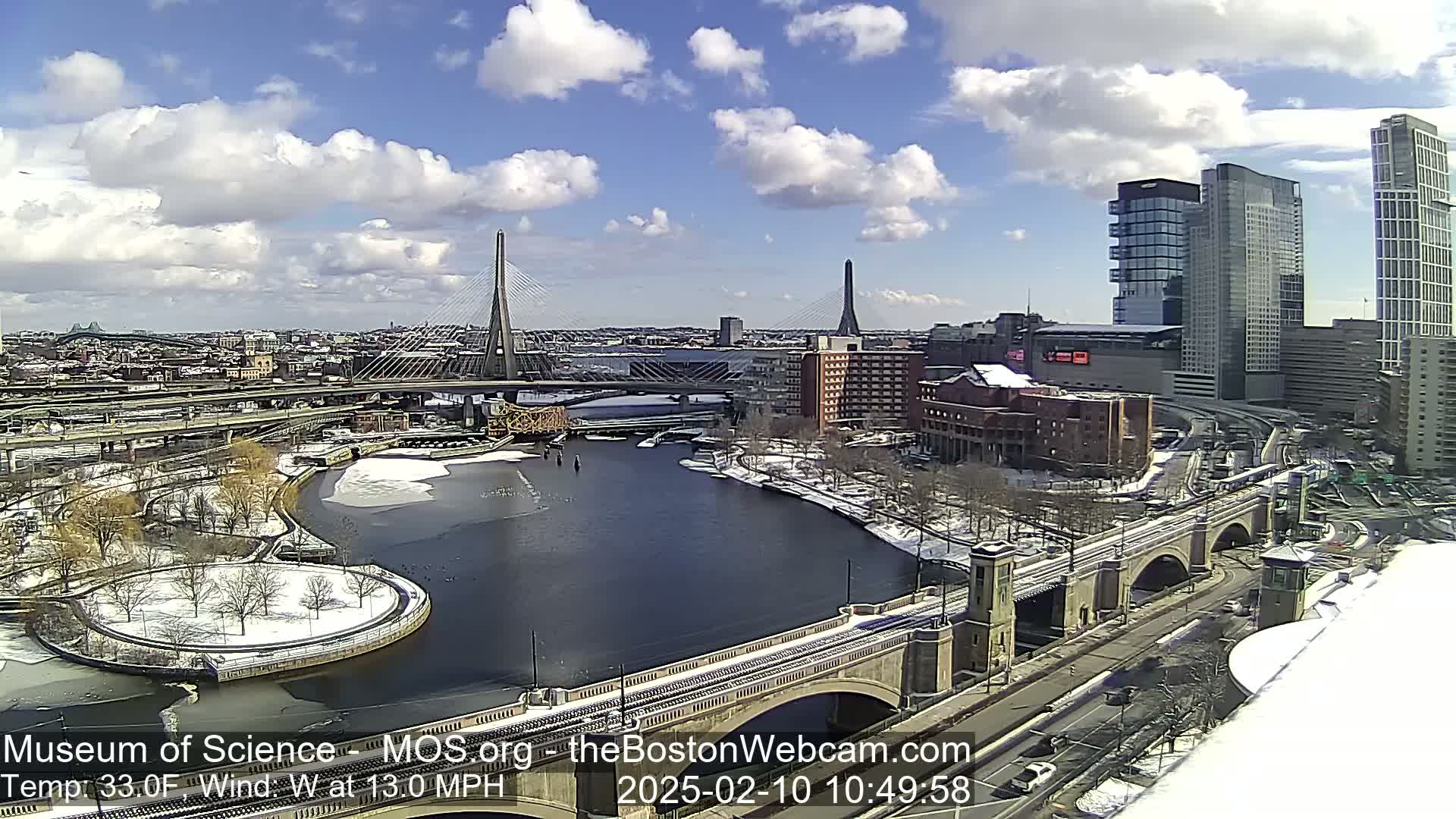 A partly cloudy day with snow on the ground shows a cityscape with two cable-stayed bridges, a river, and several buildings.