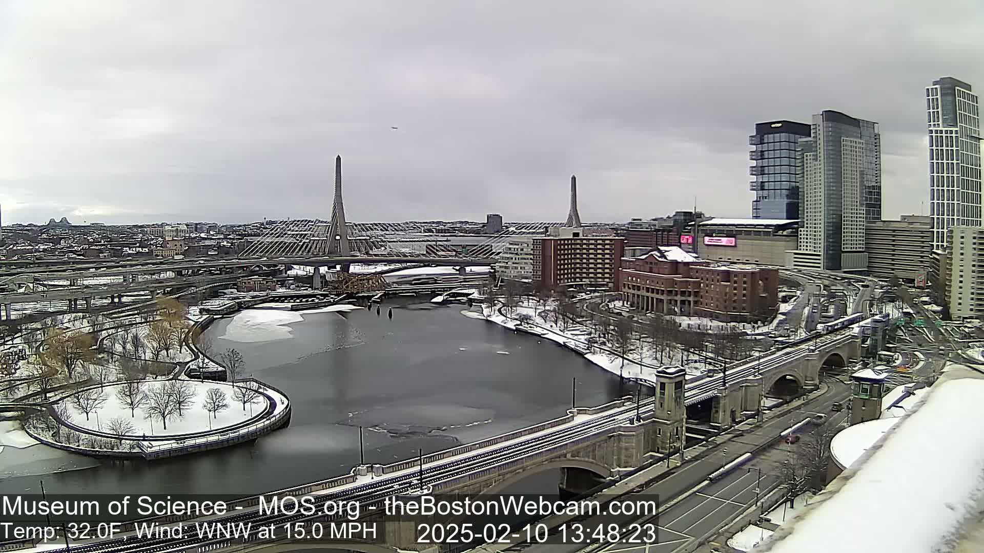 A snow-covered city scene shows a river with partially frozen water, bridges, and tall buildings under an overcast sky.