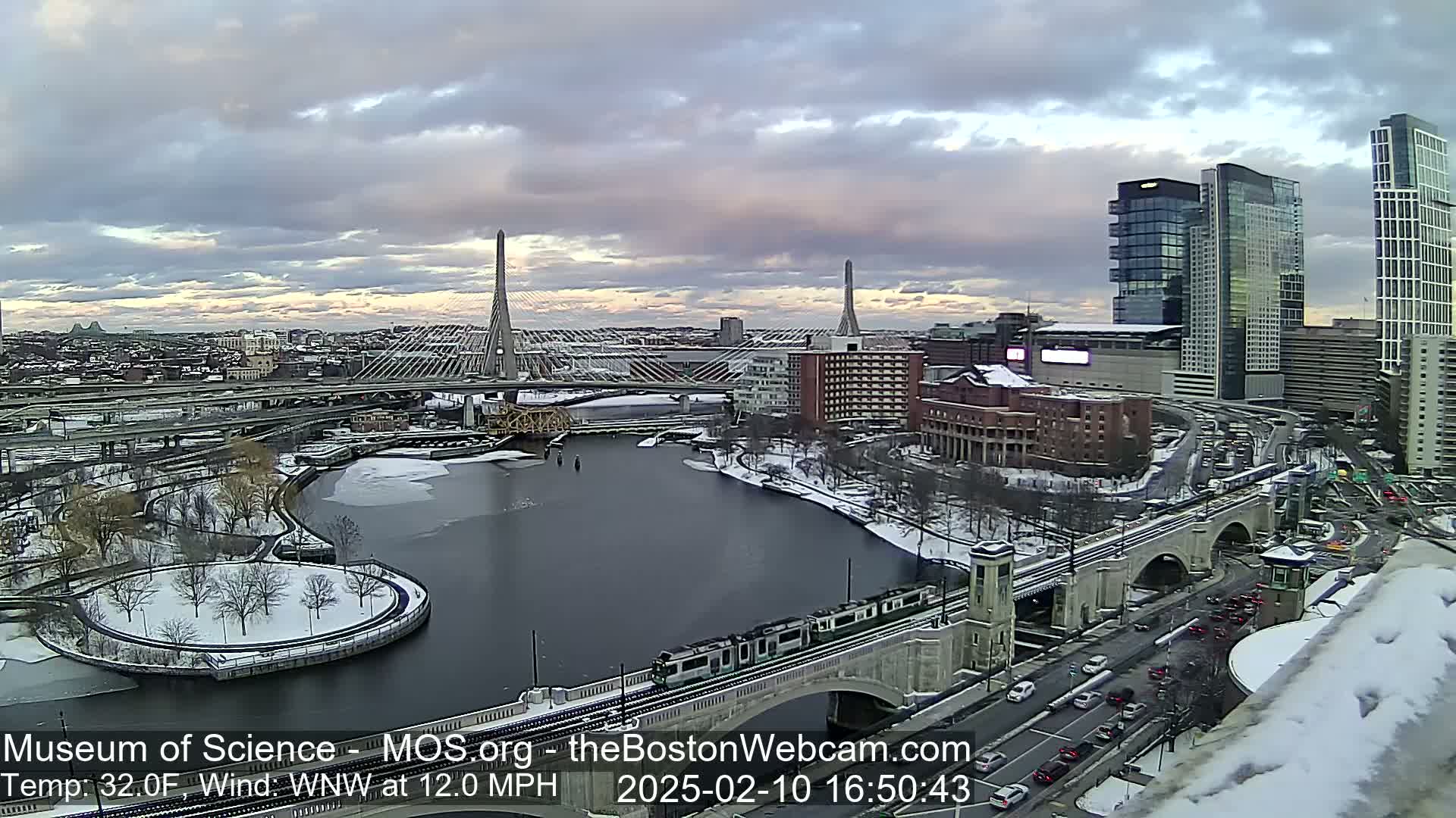 A snow-covered city landscape with two cable-stayed bridges spanning a partially frozen river under a cloudy sky.