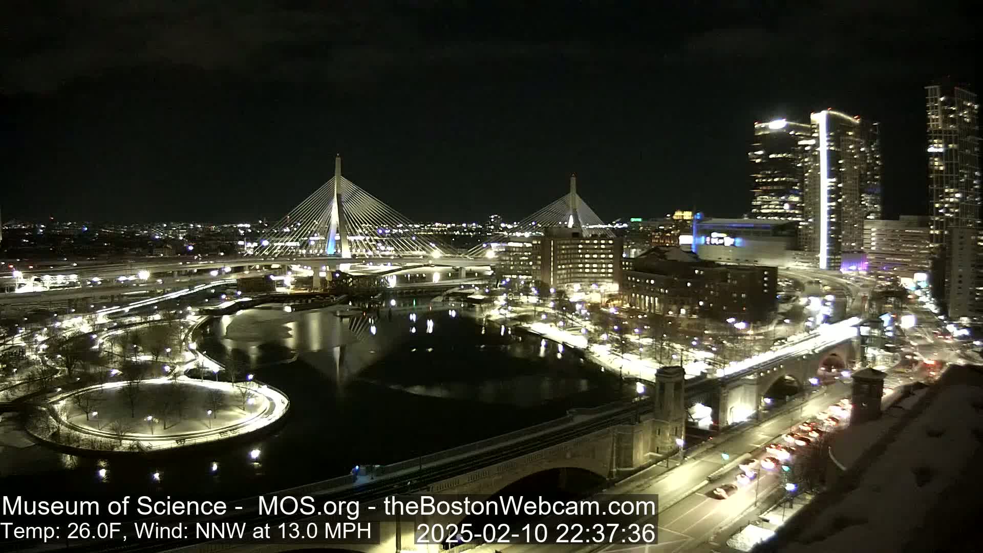 A nighttime cityscape with two cable-stayed bridges, a river, and snow-covered ground under a clear sky.