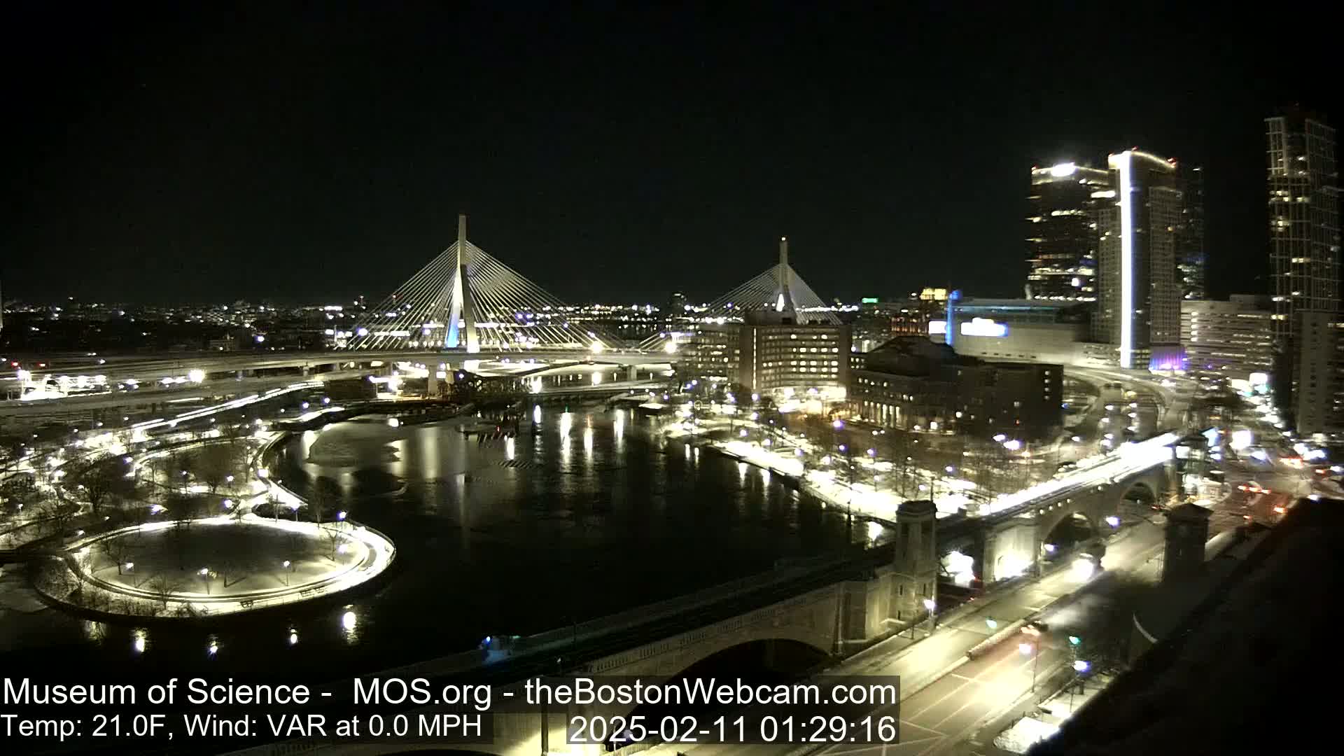 A nighttime cityscape with two illuminated cable-stayed bridges spanning a river, surrounded by city lights and snow-covered ground at 21°F with no wind.
