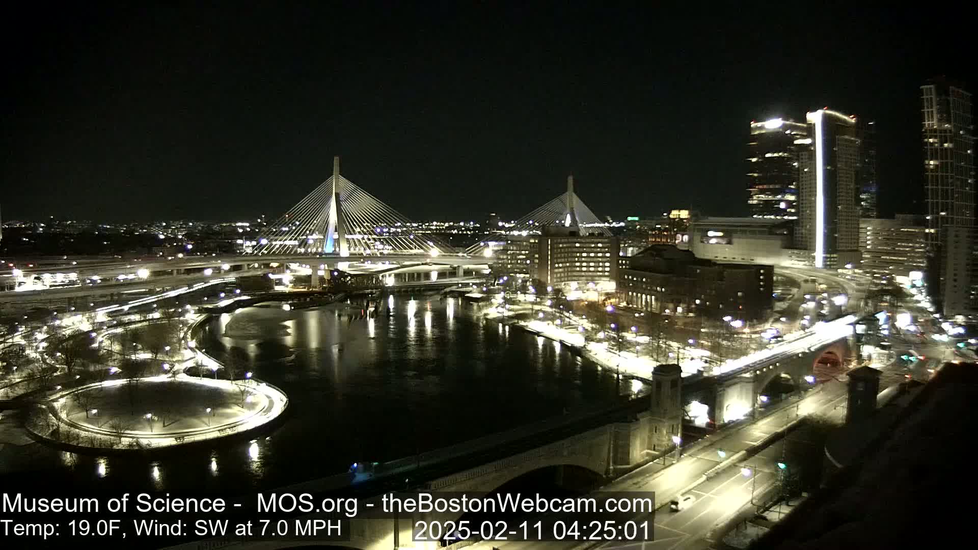 A nighttime view of two cable-stayed bridges spanning a river, with city lights and snow-covered ground, under a clear sky.