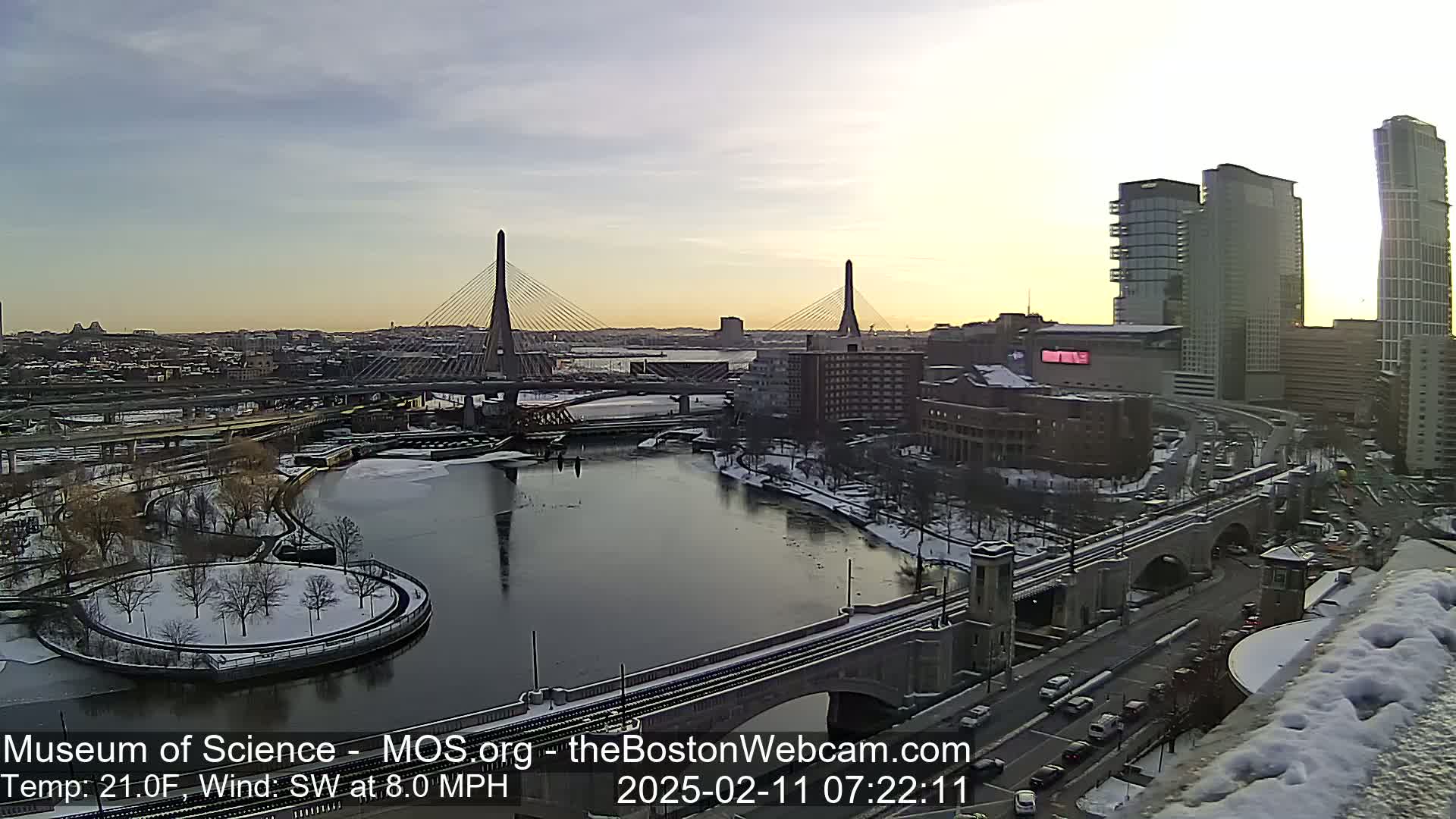 A snow-covered city scene with two cable-stayed bridges spanning a river, under a partly cloudy sky at sunrise.