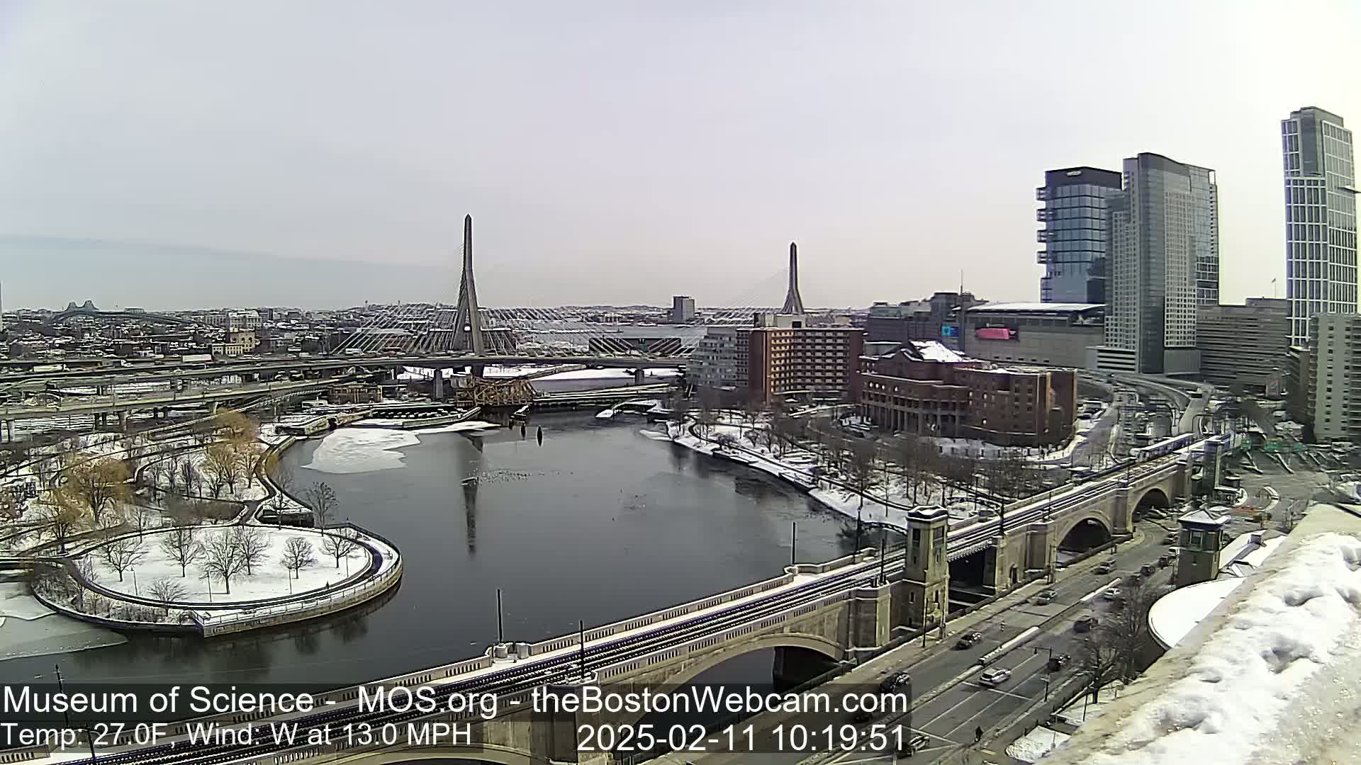 A snowy cityscape with two cable-stayed bridges spanning a river, partially frozen, under a cloudy sky.