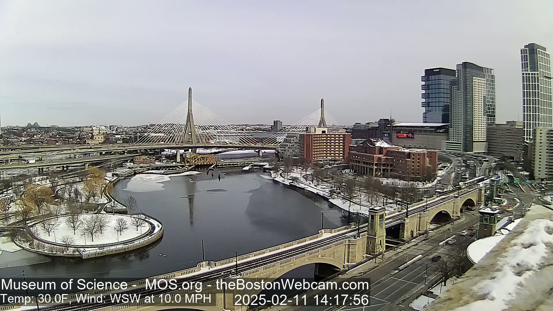 A snow-dusted cityscape with a long bridge and river, under a cloudy sky.