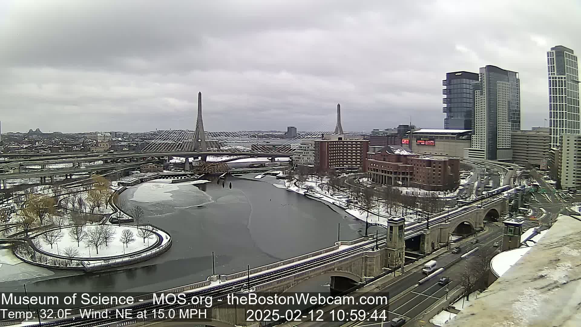 A snow-covered cityscape with a partially frozen river, two cable-stayed bridges, and high-rise buildings under an overcast sky.