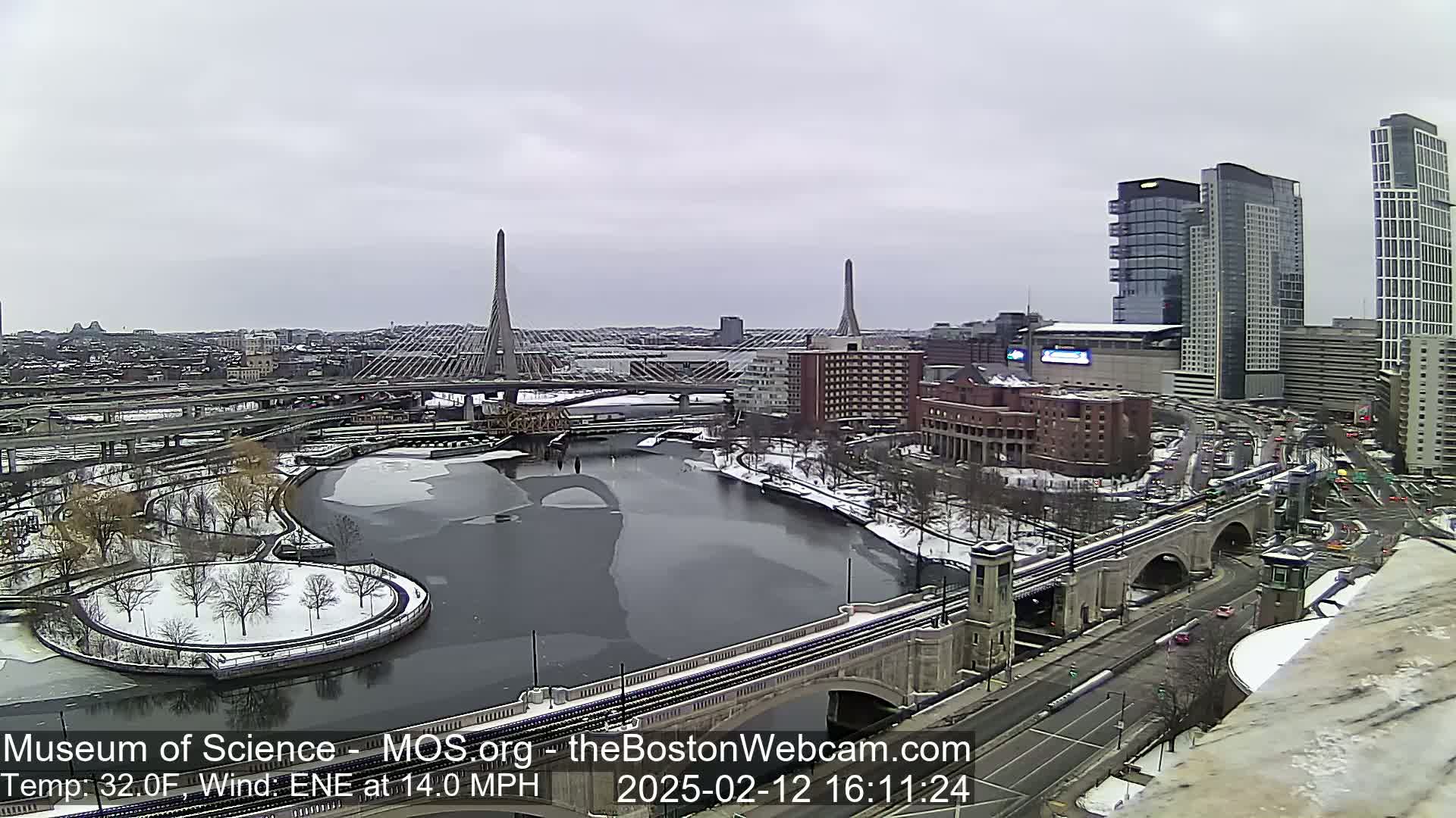 A snow-covered urban landscape with a river partially frozen, two cable-stayed bridges, and several tall buildings under an overcast sky.