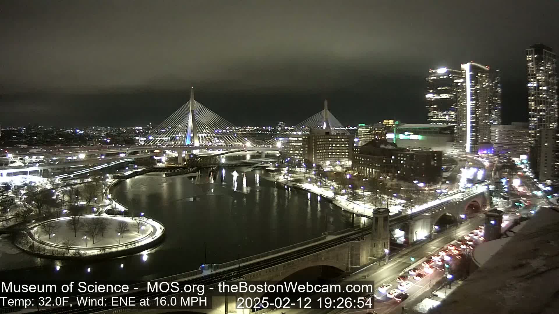 A nighttime view of a city with two cable-stayed bridges over a river, snow on the ground, and a partly cloudy sky.