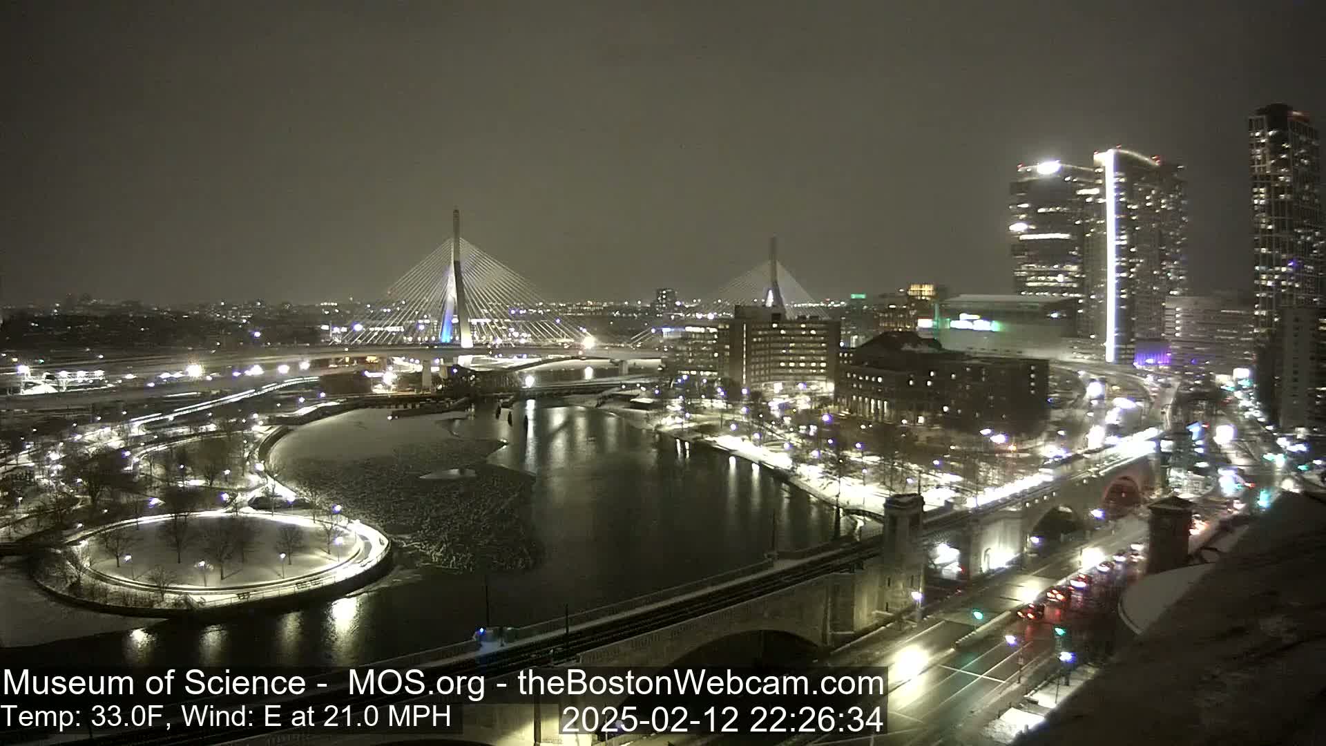 A nighttime, snowy city scene shows two cable-stayed bridges illuminated, spanning a partially frozen river, with city lights and buildings reflecting in the water.