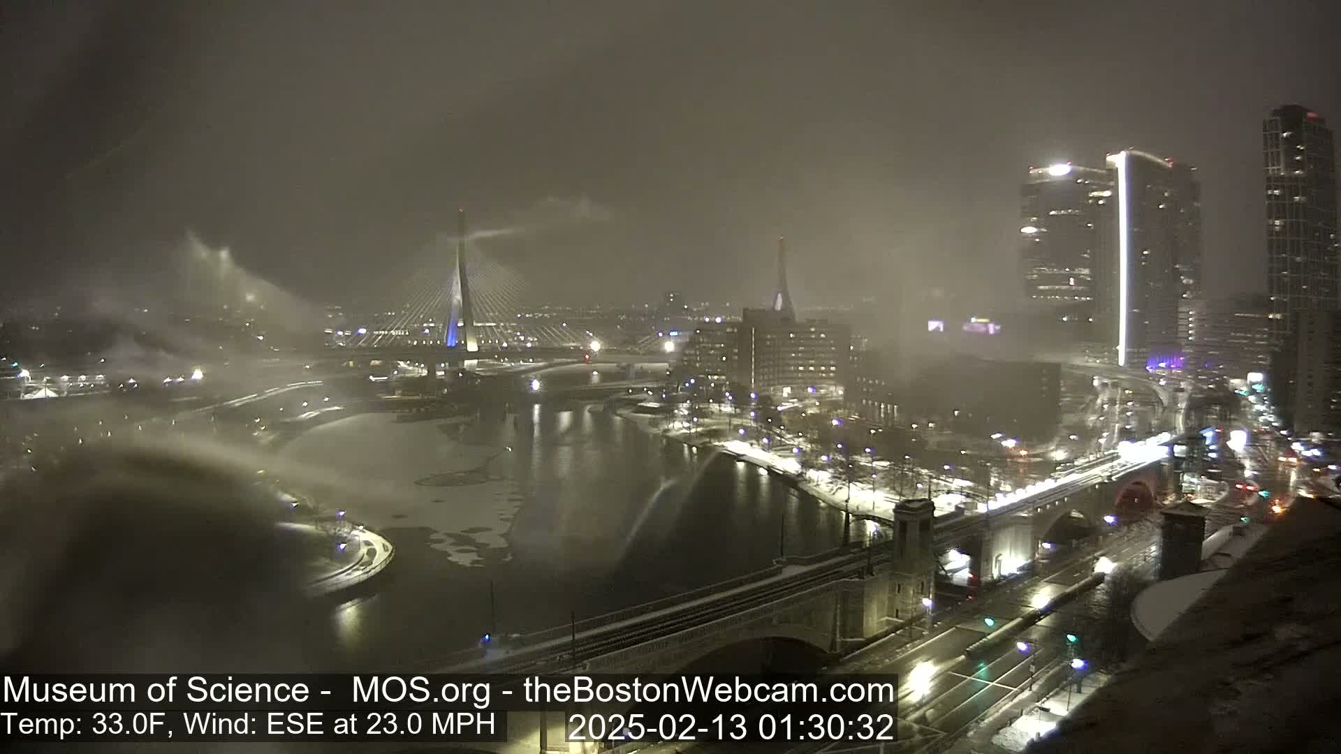 A nighttime cityscape with snow falling heavily, showing bridges and buildings along a river.