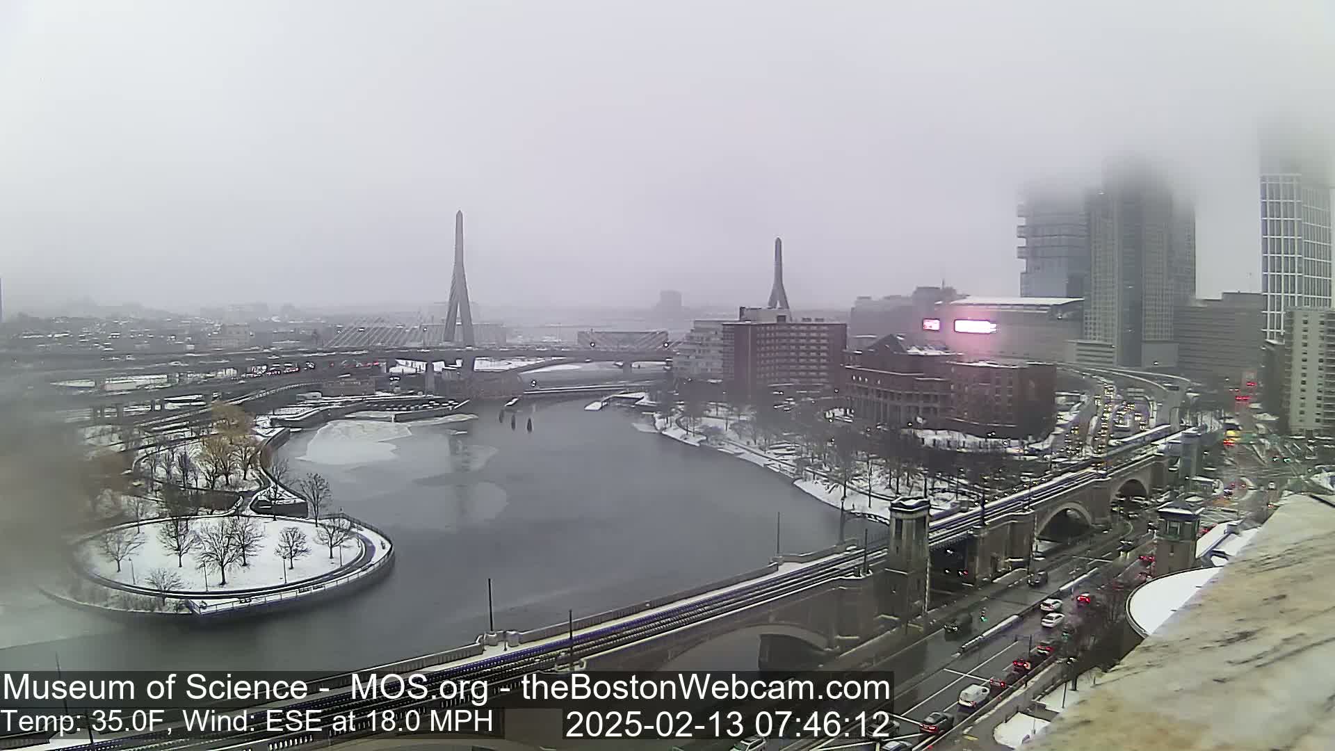 A snowy cityscape is viewed from a high vantage point, showing a river partially frozen over, bridges, and buildings under an overcast sky with an 18 mph wind.