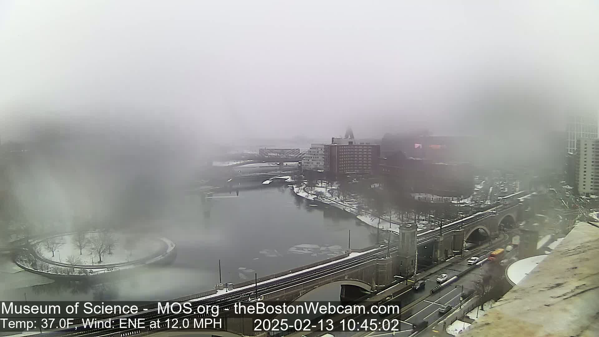 A foggy, snowy cityscape is viewed from above, showing a river with ice floes, a bridge with vehicles, and buildings partially obscured by the thick fog.