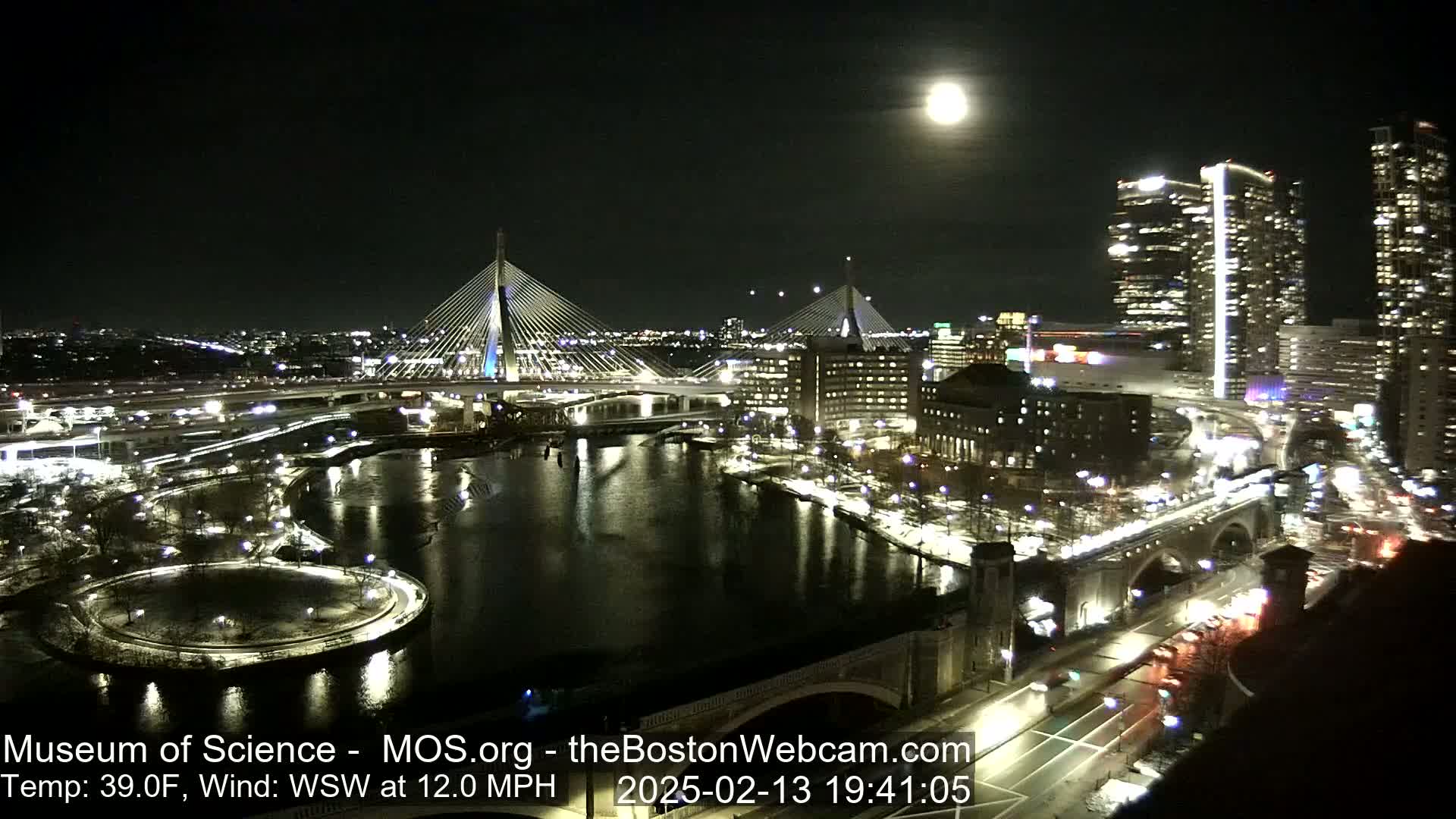 A nighttime view of a city with two cable-stayed bridges spanning a river, under a bright full moon and a 12 mph wind, with a temperature of 39°F.