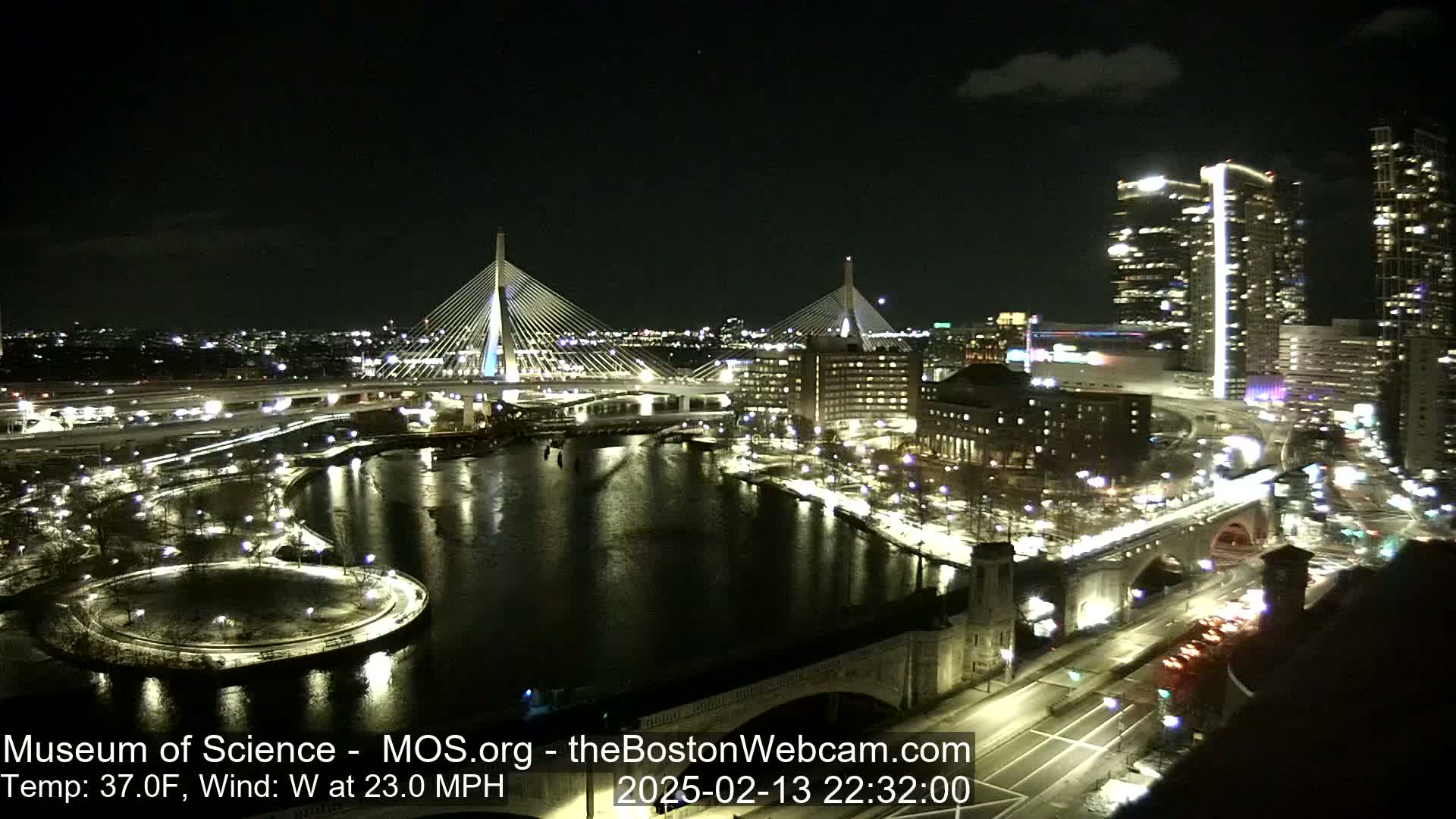 A nighttime cityscape with two illuminated cable-stayed bridges spanning a dark river, surrounded by city lights and buildings, under a clear sky with a temperature of 37°F and a 23 mph west wind.