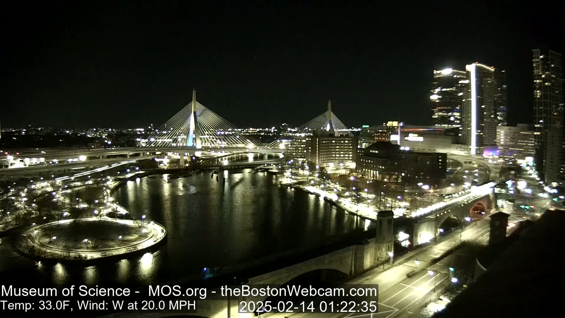 A nighttime cityscape with two illuminated cable-stayed bridges spanning a dark river, surrounded by city lights and some snow on the ground, under a clear sky with a 33°F temperature and a 20 mph west wind.