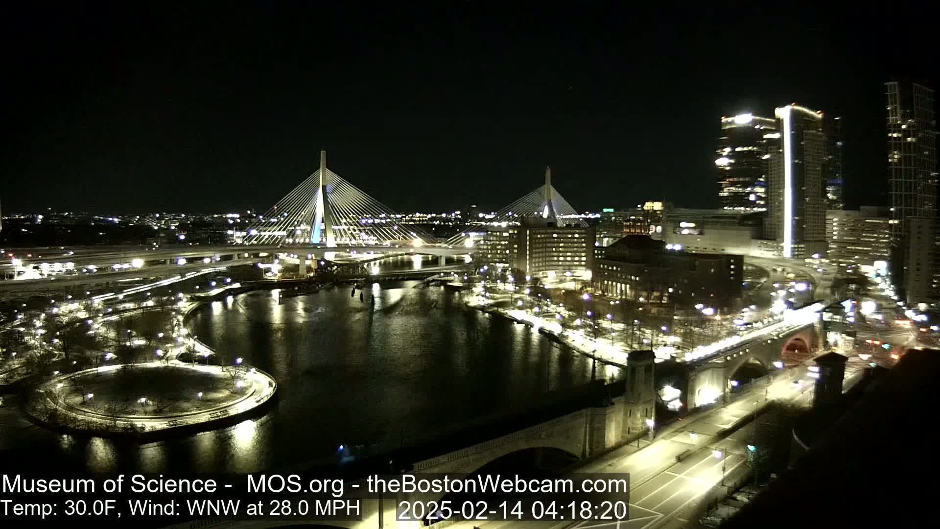A nighttime aerial view of a city with two illuminated cable-stayed bridges spanning a river, surrounded by buildings and roadways, with a WNW wind at 28 mph and a temperature of 30°F.