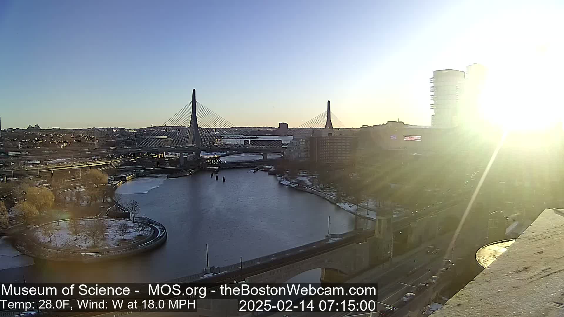 A sunny, cold morning view of a city with two cable-stayed bridges spanning a partially frozen river, with some snow visible on the ground.