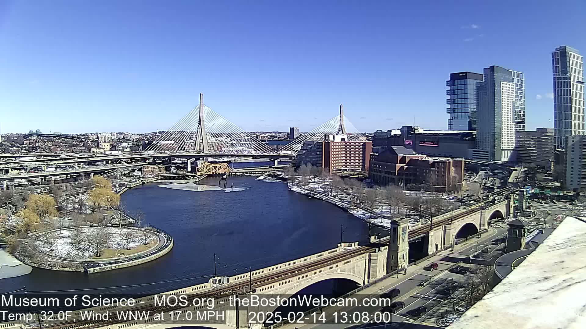 A sunny, cold day with a 17 mph wind shows a city scene with two cable-stayed bridges spanning a river, with some snow visible on the ground and trees.