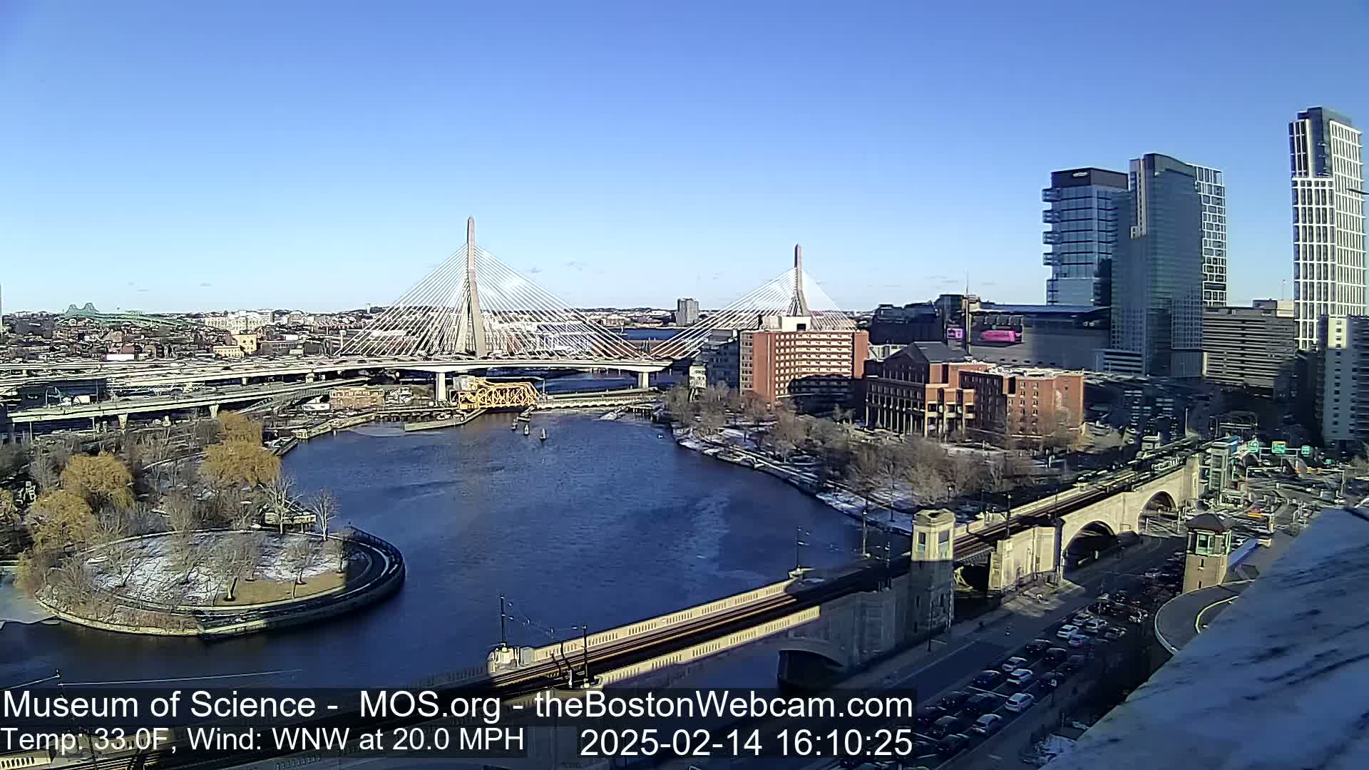 A sunny day with a 20 mph wind reveals a cityscape featuring a river, two cable-stayed bridges, and several modern high-rise buildings, some with patches of snow on the ground.