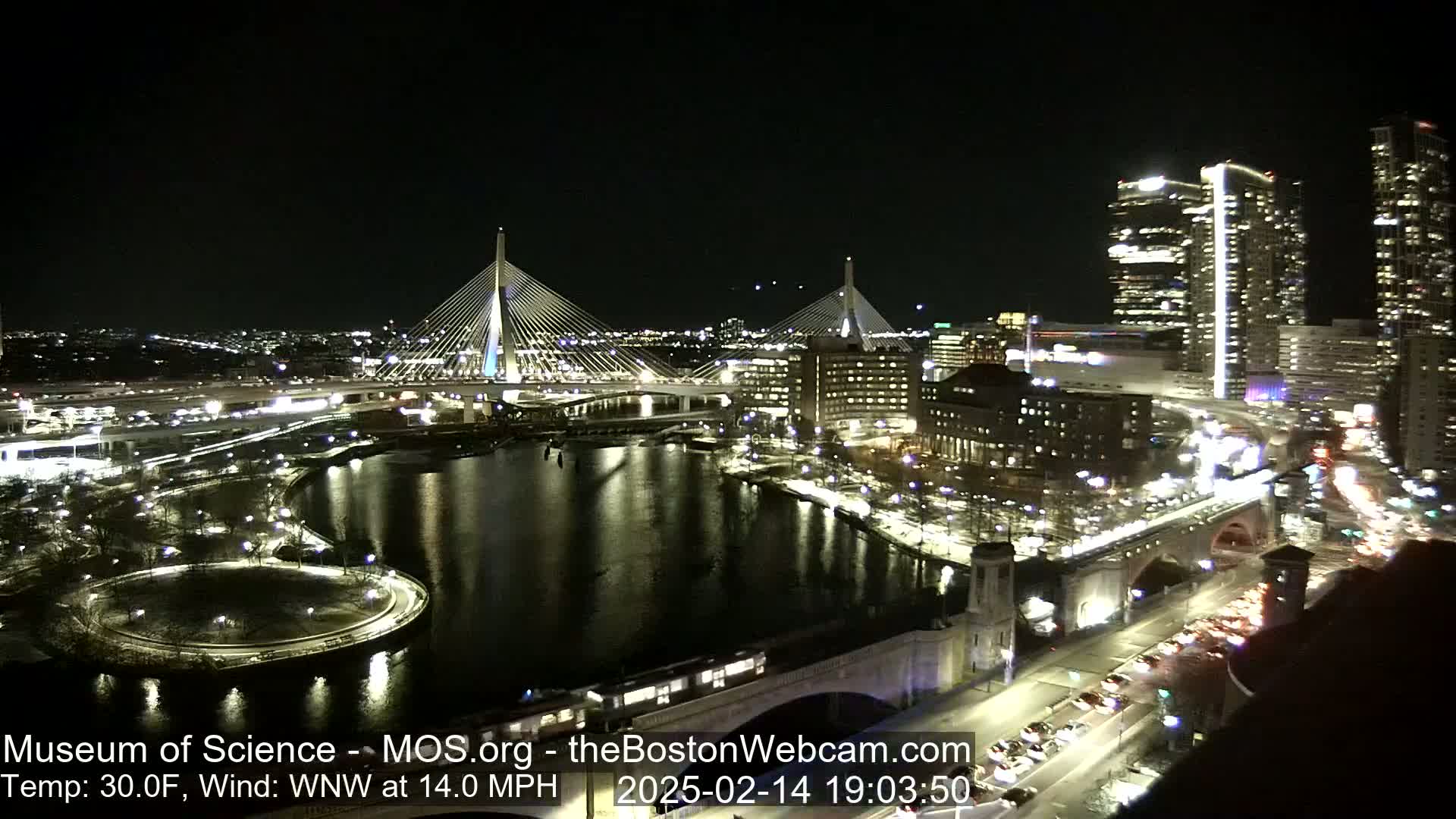 A nighttime aerial view of a city with two illuminated cable-stayed bridges spanning a dark river, surrounded by brightly lit buildings and roads with traffic, under a clear sky.