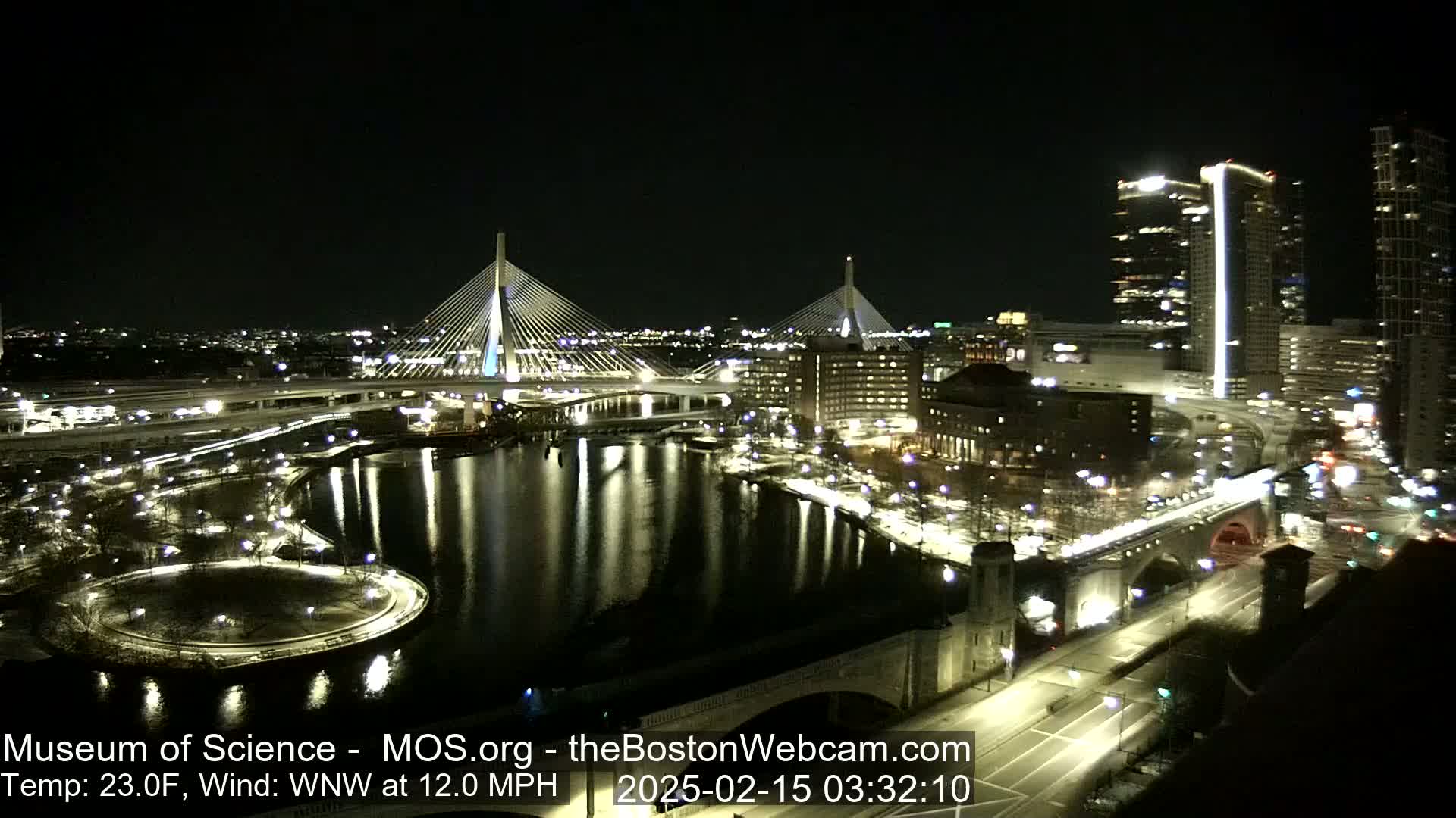A nighttime view of a city illuminated with bridges, waterways, and buildings, with a wind from the west-northwest at 12 mph and a temperature of 23 degrees Fahrenheit.