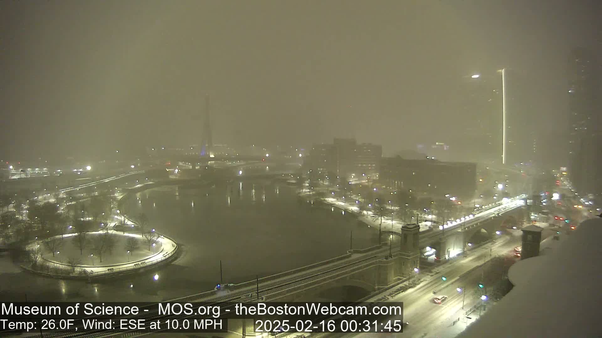A snow-covered cityscape at night is viewed from above, showing a river, a bridge, and city lights obscured by heavy snowfall with a 10 mph wind.