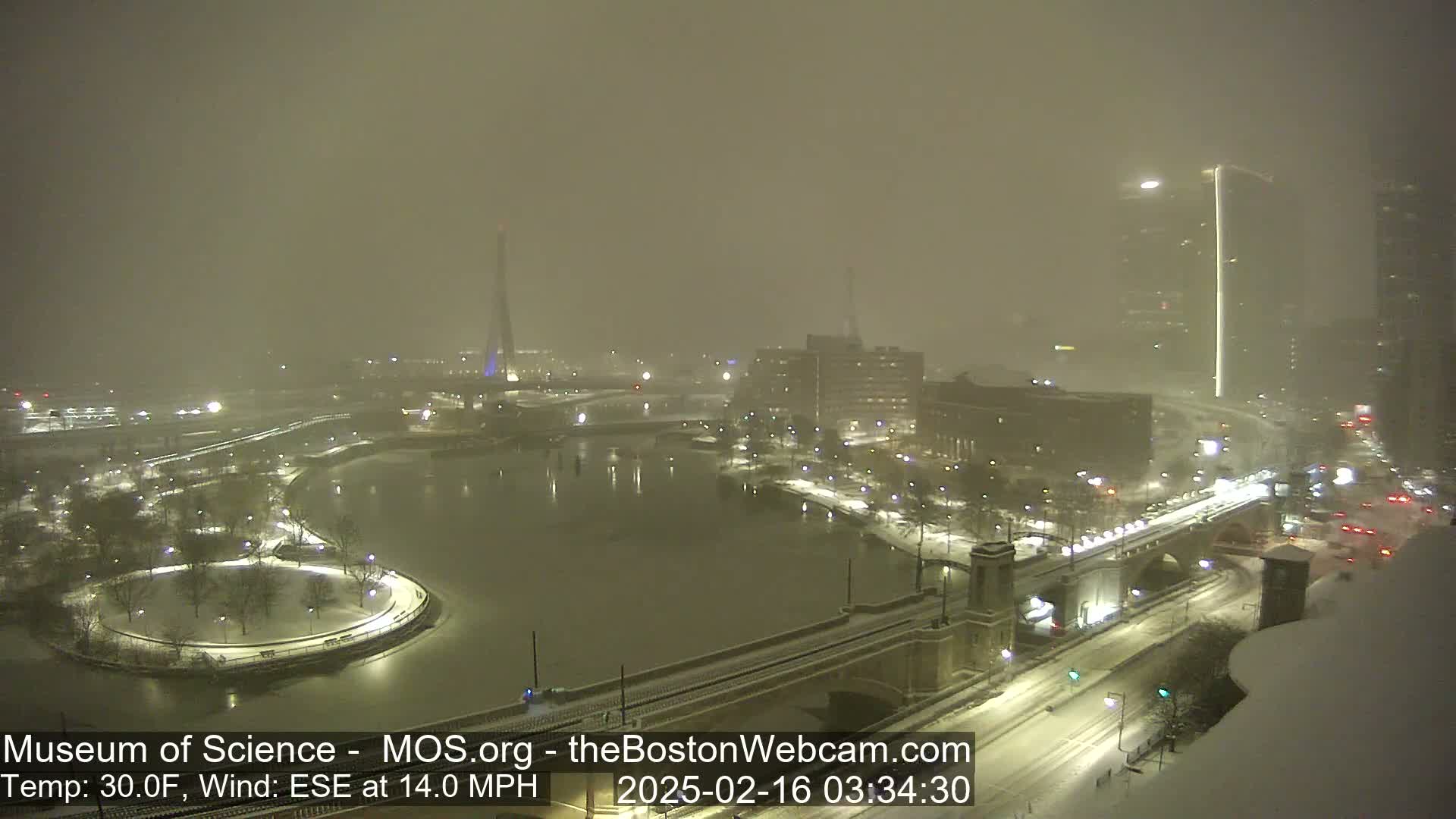 A snow-covered cityscape at night, viewed from above, showing a river, bridges, and buildings under heavy snowfall with a 30°F temperature and 14 mph wind from the east-southeast.