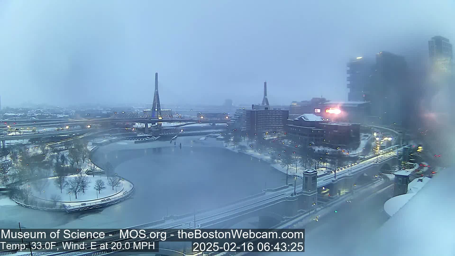 A snowy cityscape with two tall towers and a bridge, viewed from a high vantage point in windy, 33°F weather.