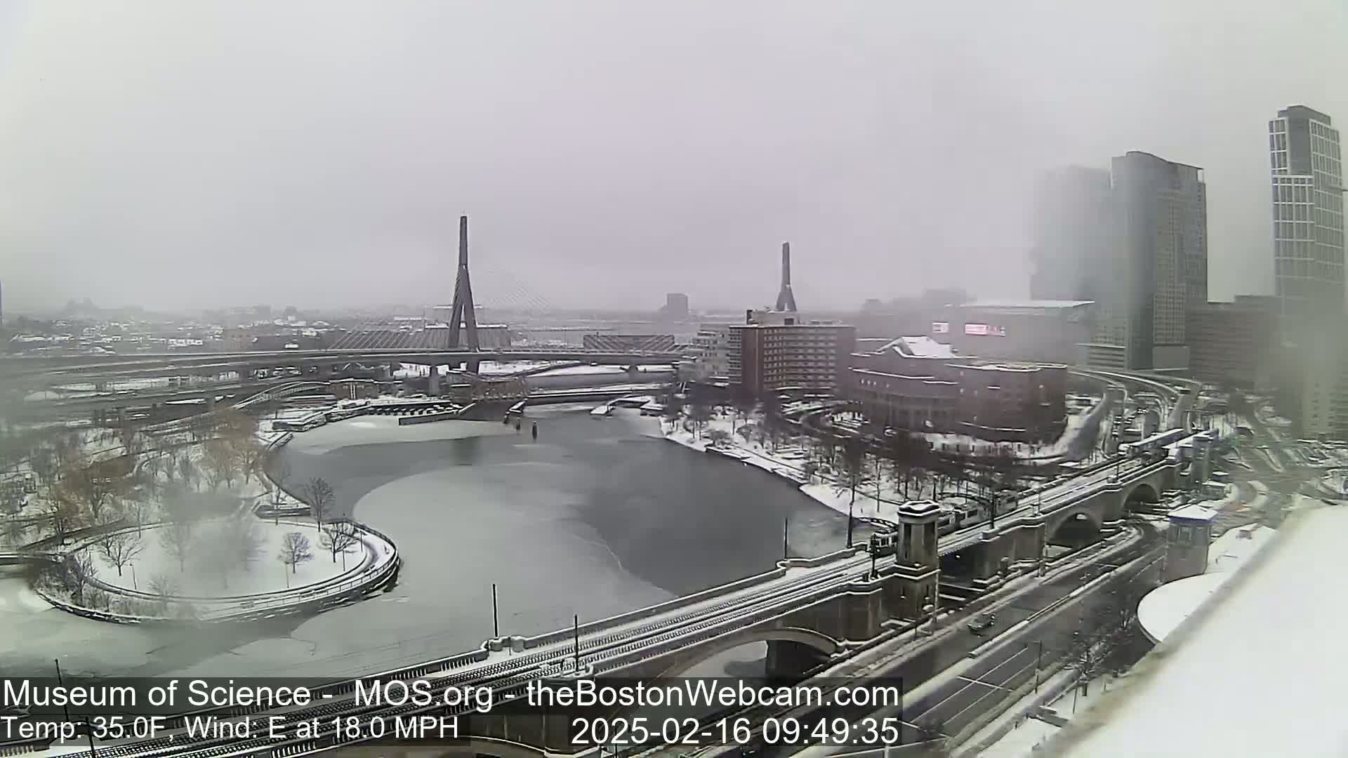 A snow-covered urban landscape with two cable-stayed bridges and a partially frozen river, under a heavy snowfall with an easterly wind.