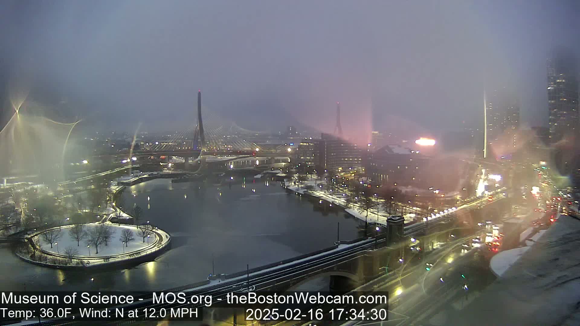 A snowy cityscape at night, viewed from an elevated position, shows a bridge over a partially frozen river with city lights and traffic visible under a hazy sky.