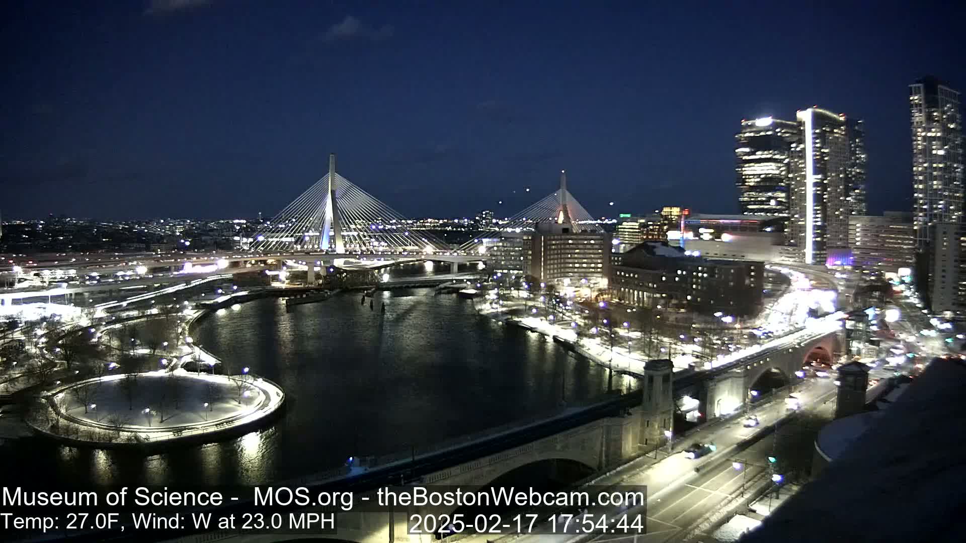 A nighttime view of a city with two cable-stayed bridges spanning a river, under a clear, dark sky, with snow on the ground and a 27°F temperature and 23 mph west wind.