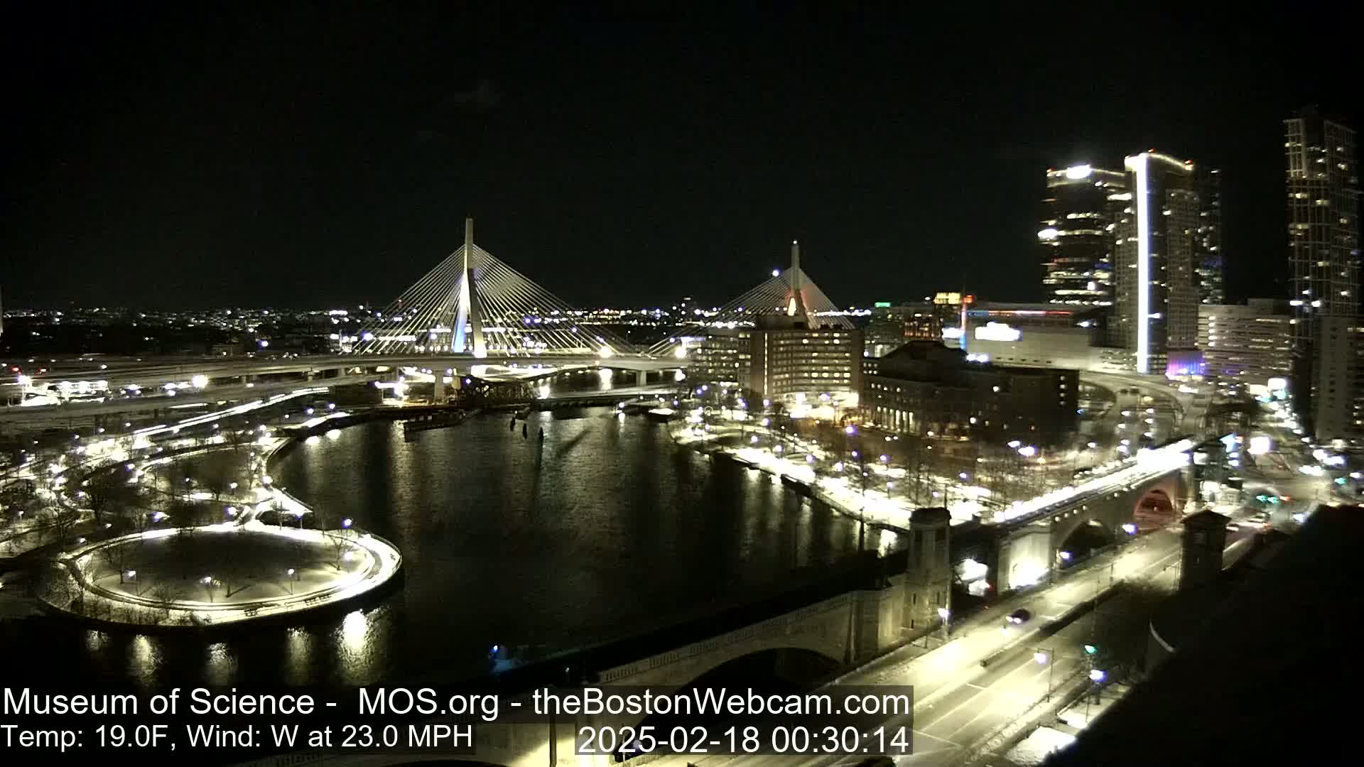 A nighttime cityscape showing two cable-stayed bridges over a dark river, with city lights illuminating snow-covered ground and roads, under a clear sky and a 19°F temperature with a 23 mph west wind.