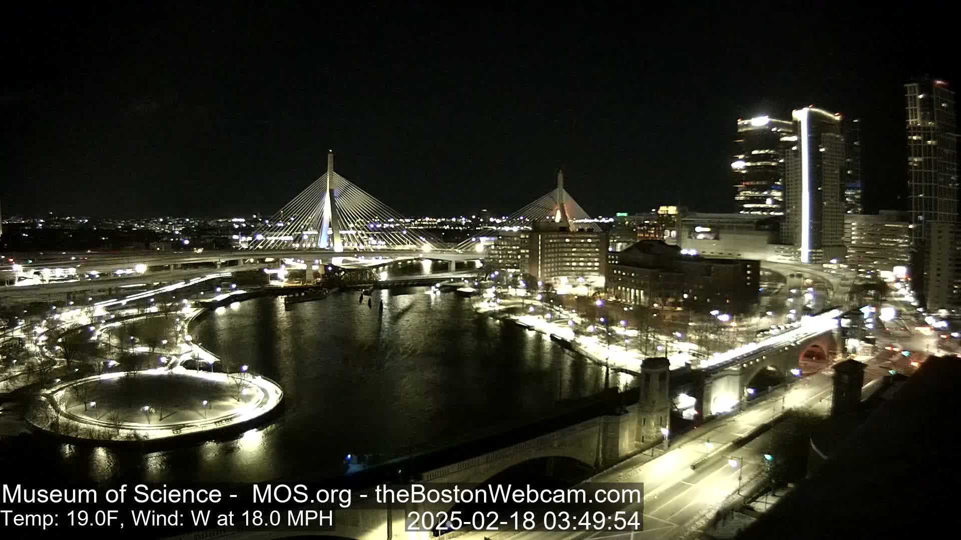 A nighttime cityscape featuring two illuminated cable-stay bridges spanning a dark river, with city lights reflecting on the water, under a clear night sky with a westerly wind at 18 mph and a temperature of 19°F.