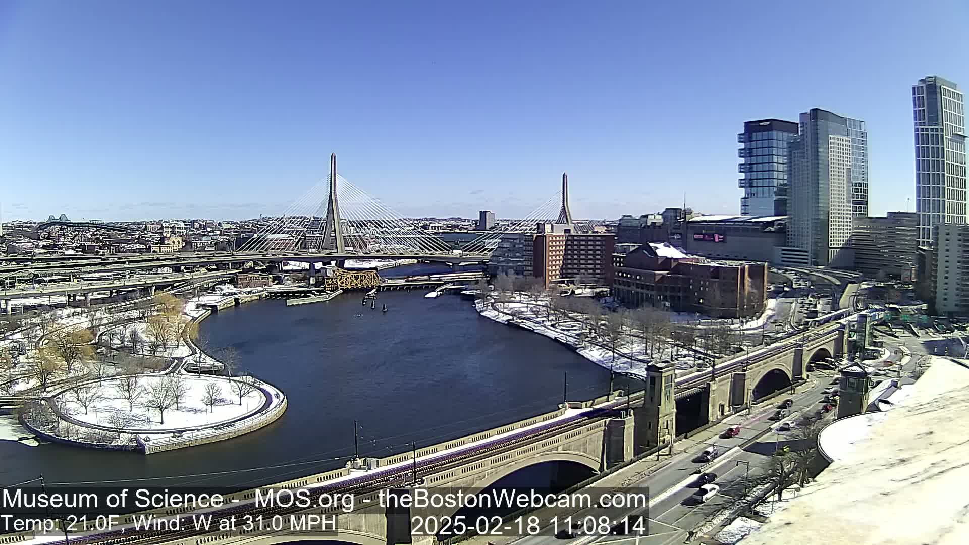 A snow-dusted cityscape with a river, bridges, and modern high-rises under a clear blue sky, with a brisk westerly wind.