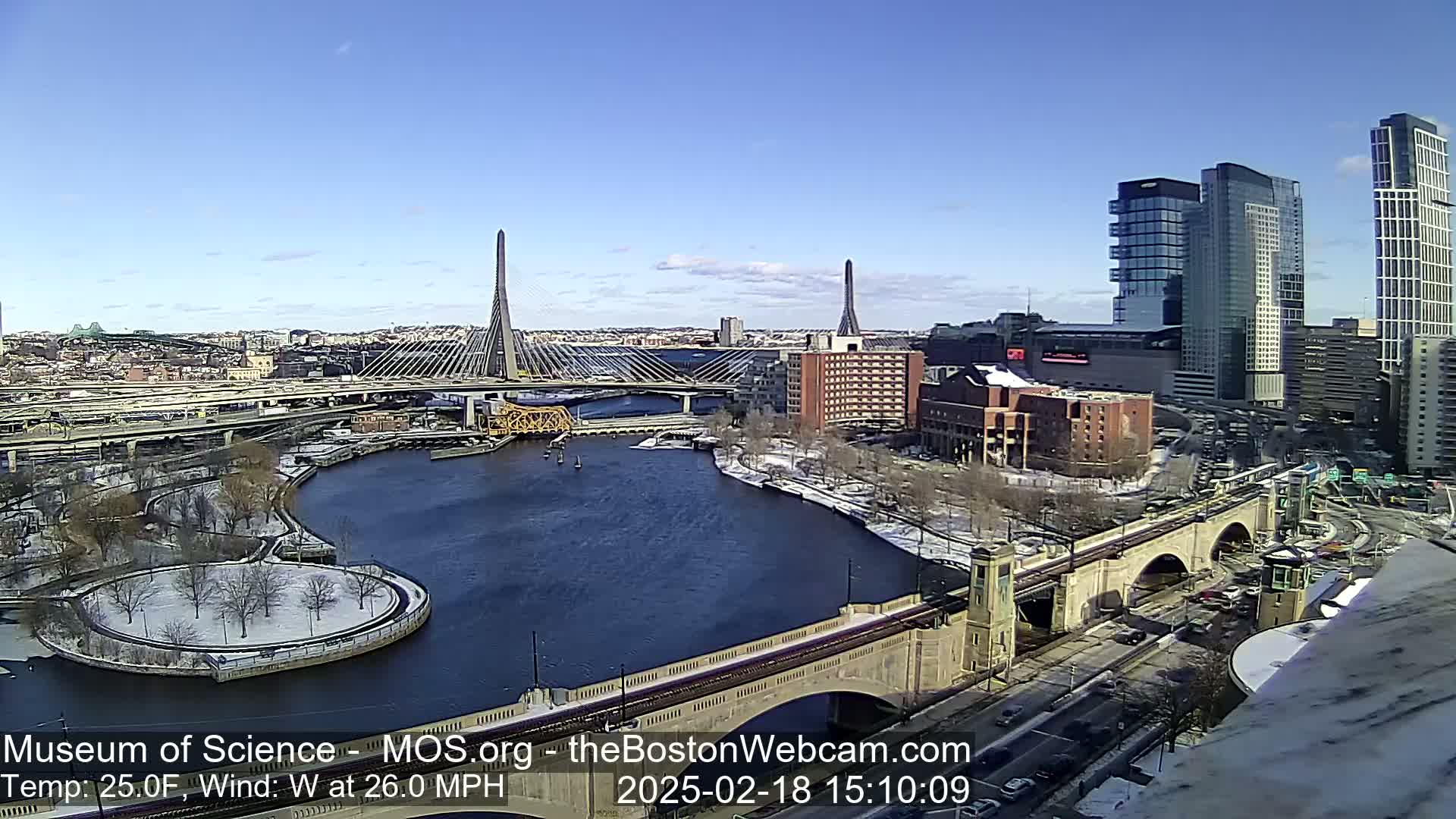A sunny day with a 26 mph west wind shows a cityscape with two cable-stayed bridges spanning a river, parts of which are iced over, with snow-covered land surrounding the waterway.