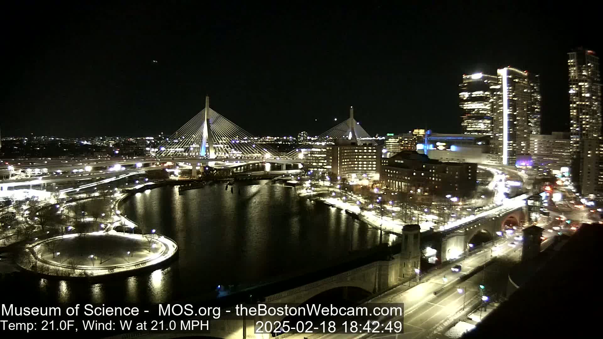 A nighttime view of a city waterfront, illuminated with lights, featuring two cable-stayed bridges and a river with snow on the ground, under a clear, dark sky with a 21°F temperature and 21 mph westerly wind.