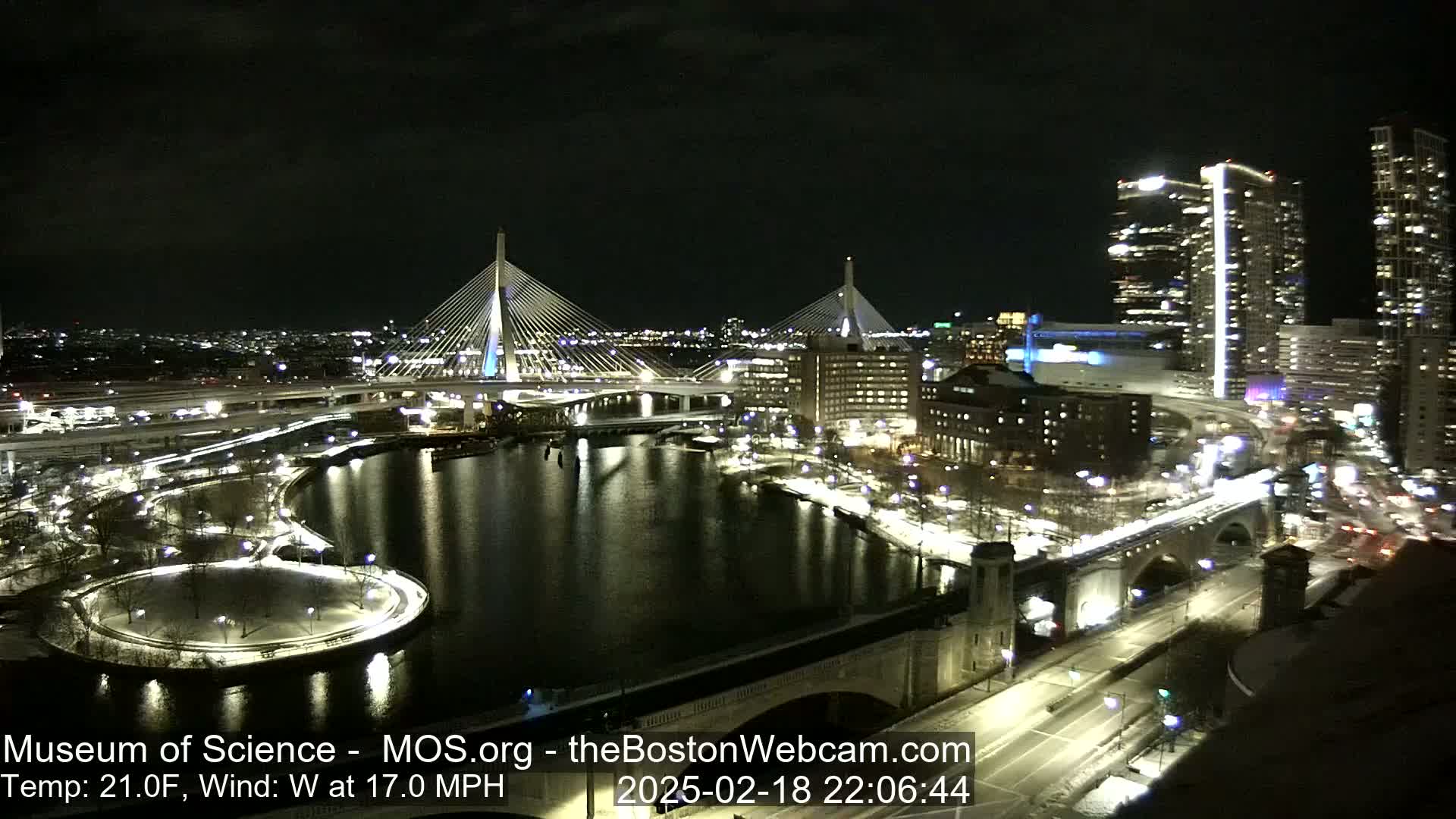 A nighttime view of a city with two illuminated cable-stayed bridges spanning a dark river, under a clear sky with a 21°F temperature and a 17 mph west wind.