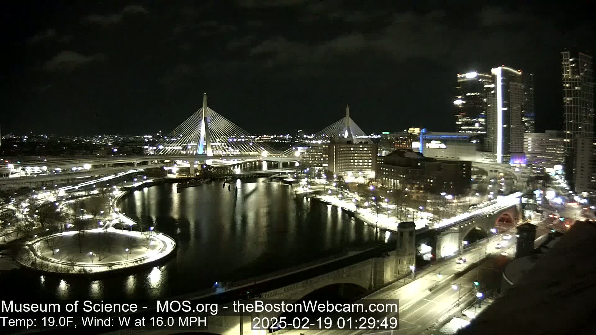 A nighttime aerial view of a city with two illuminated cable-stayed bridges spanning a dark river, surrounded by snow-covered parkland and lit-up buildings under a partly cloudy sky, with a strong west wind.