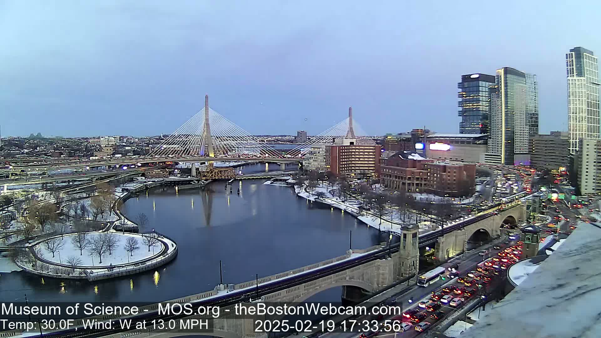 A snowy cityscape at dusk shows two cable-stayed bridges spanning a river, with heavy traffic on surrounding roads and a partially frozen river.