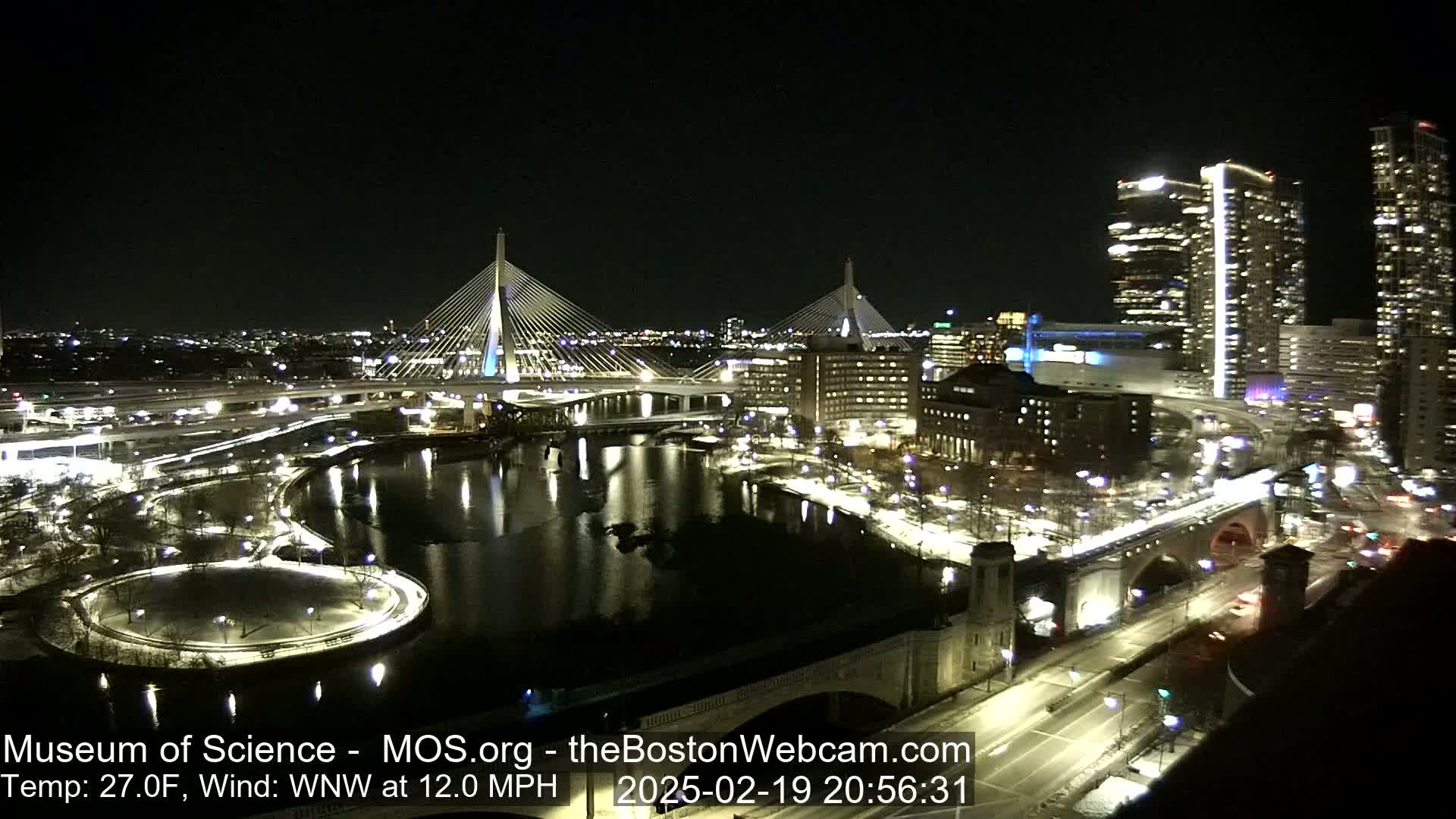 A nighttime cityscape with two illuminated cable-stayed bridges spanning a dark river, surrounded by city lights and buildings under a clear sky, with a temperature of 27 degrees Fahrenheit and a wind from the west-northwest at 12 miles per hour.