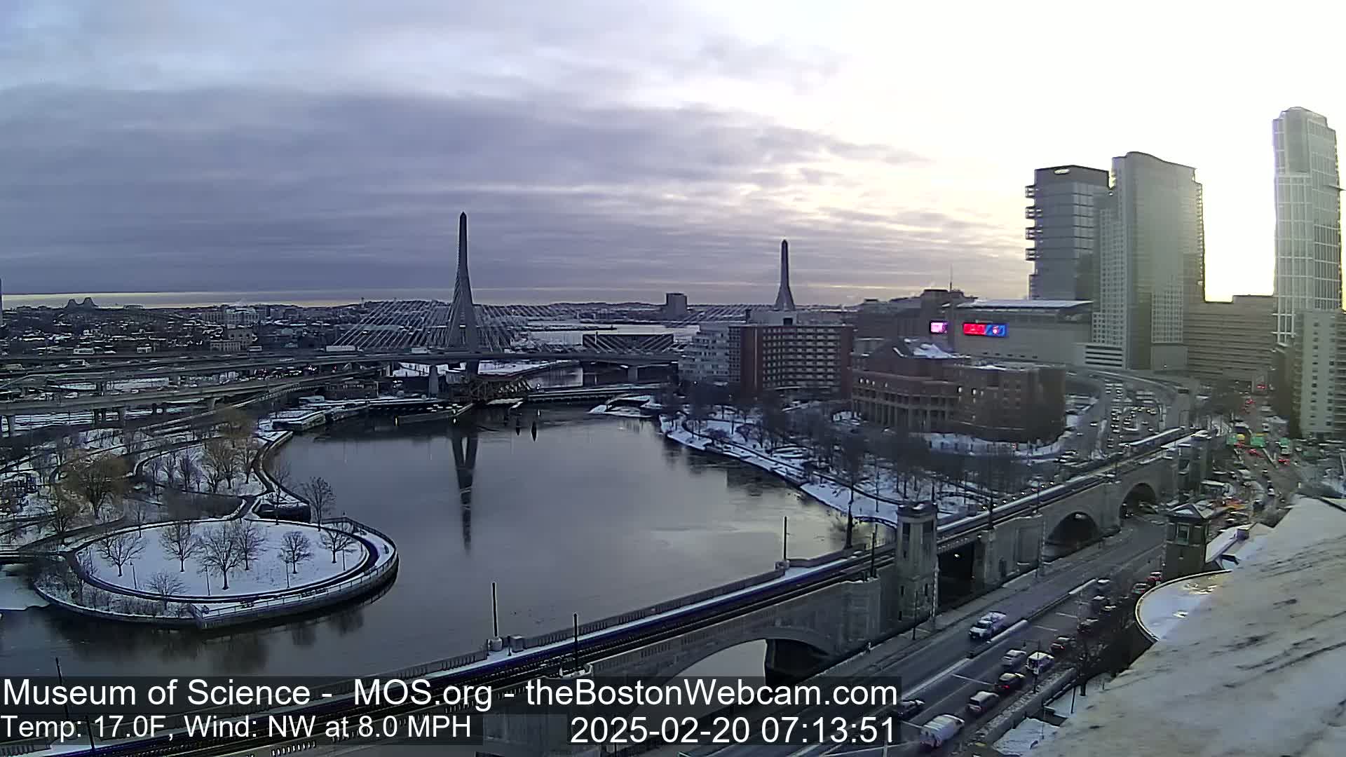 A snow-dusted cityscape with two cable-stayed bridges spanning a river, under a partly cloudy sky at sunrise.