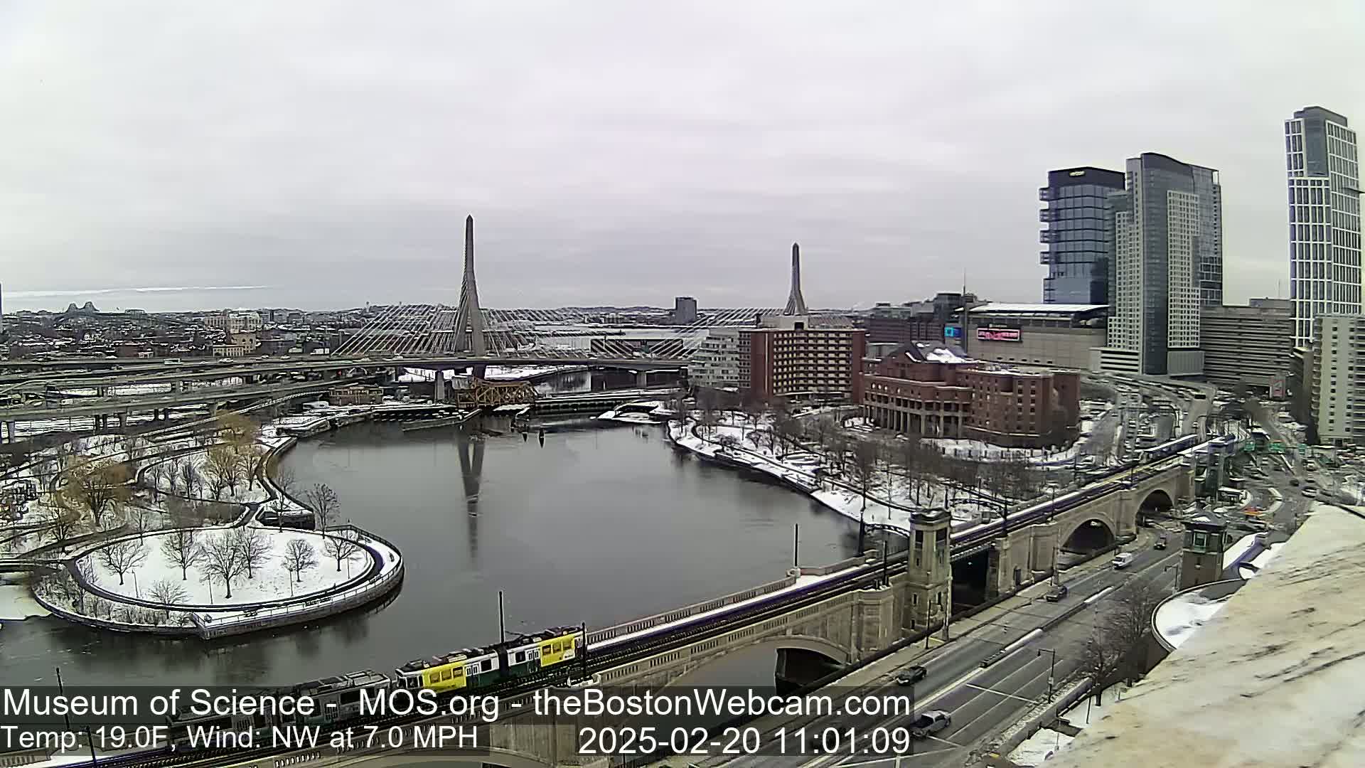 A snowy cityscape with two cable-stayed bridges spanning a river, alongside a train crossing a stone bridge, under a cloudy sky.