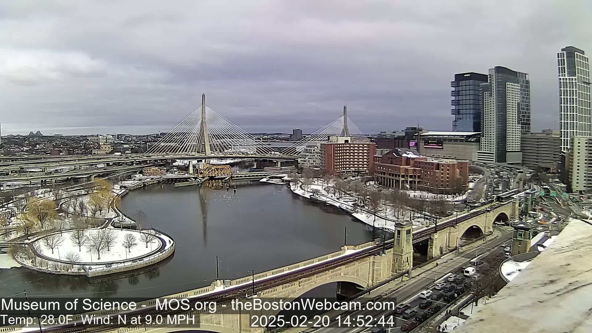 A snowy cityscape with two cable-stayed bridges spanning a river, under an overcast sky.