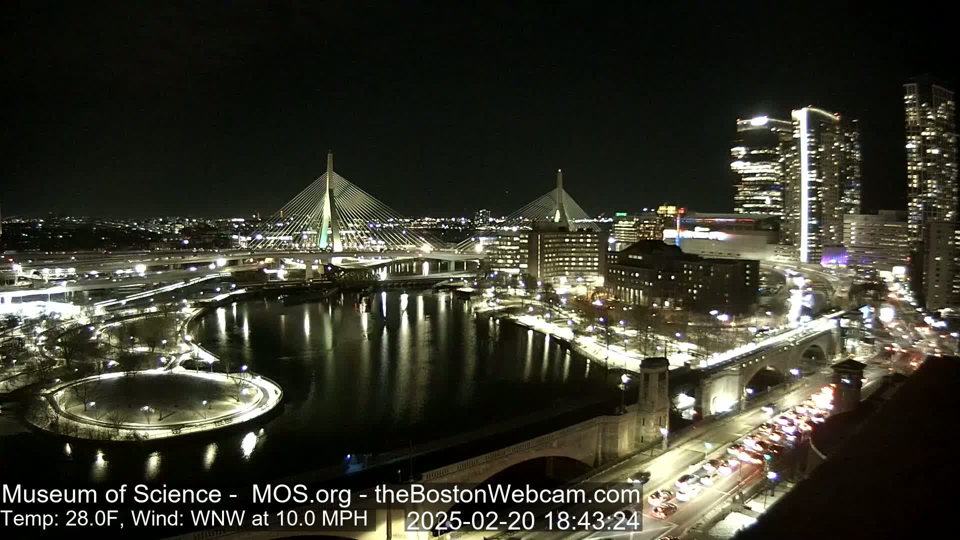 A nighttime view of two illuminated cable-stayed bridges spanning a river, with city lights reflecting in the water, under a clear, cold sky.