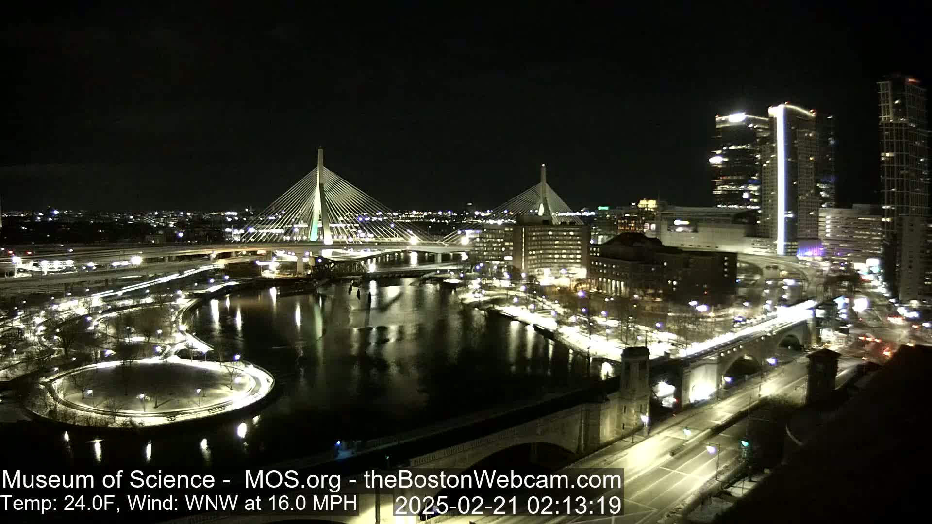 A nighttime aerial view of a city with two cable-stayed bridges, a river, and snow-covered ground, under a clear sky with a temperature of 24°F and a 16 mph wind from the west-northwest.