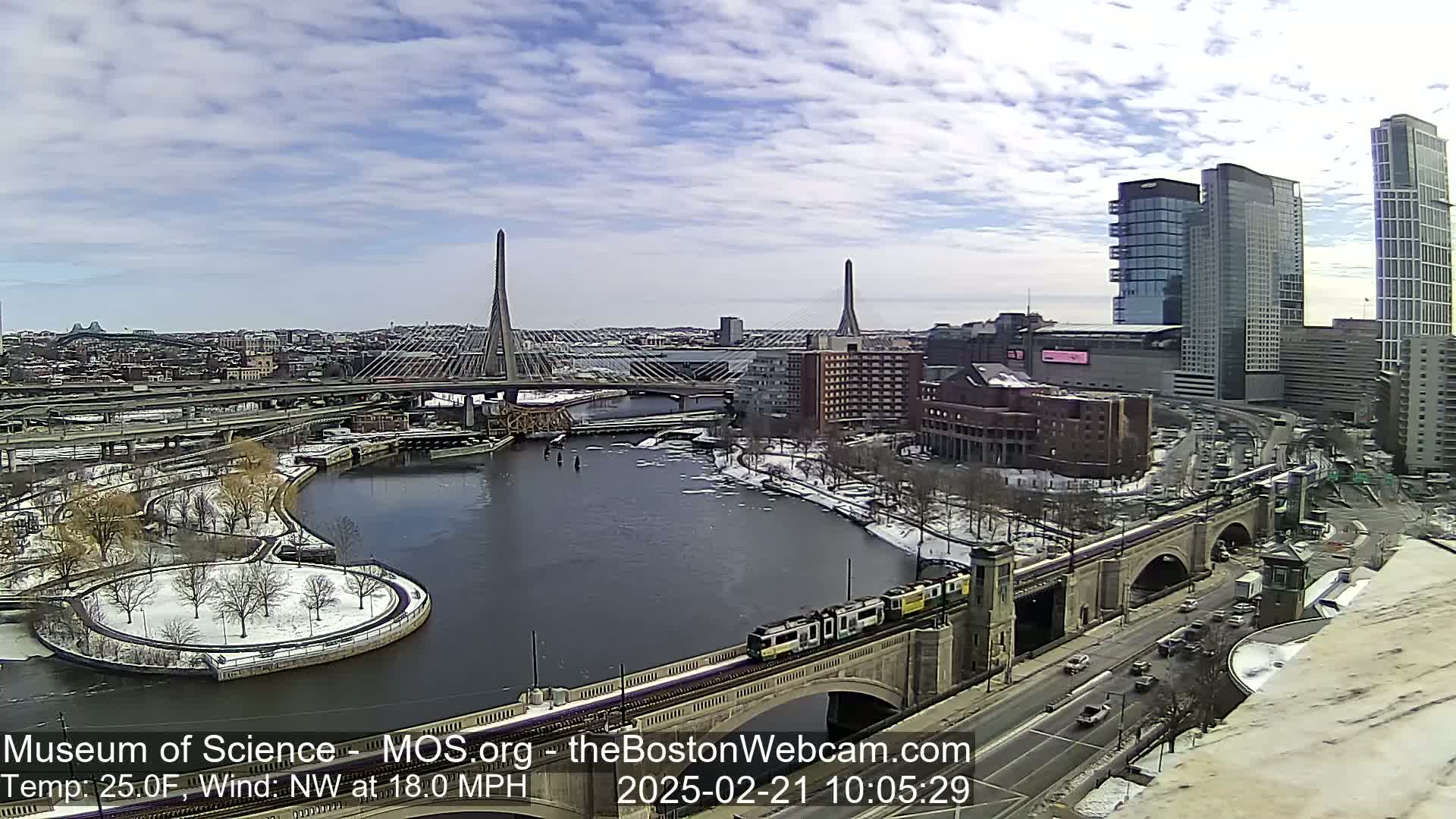 A partly cloudy day with a northwest wind at 18 mph and a temperature of 25°F shows a city scene with two cable-stayed bridges, a river, a train crossing a bridge, and snow-covered ground.