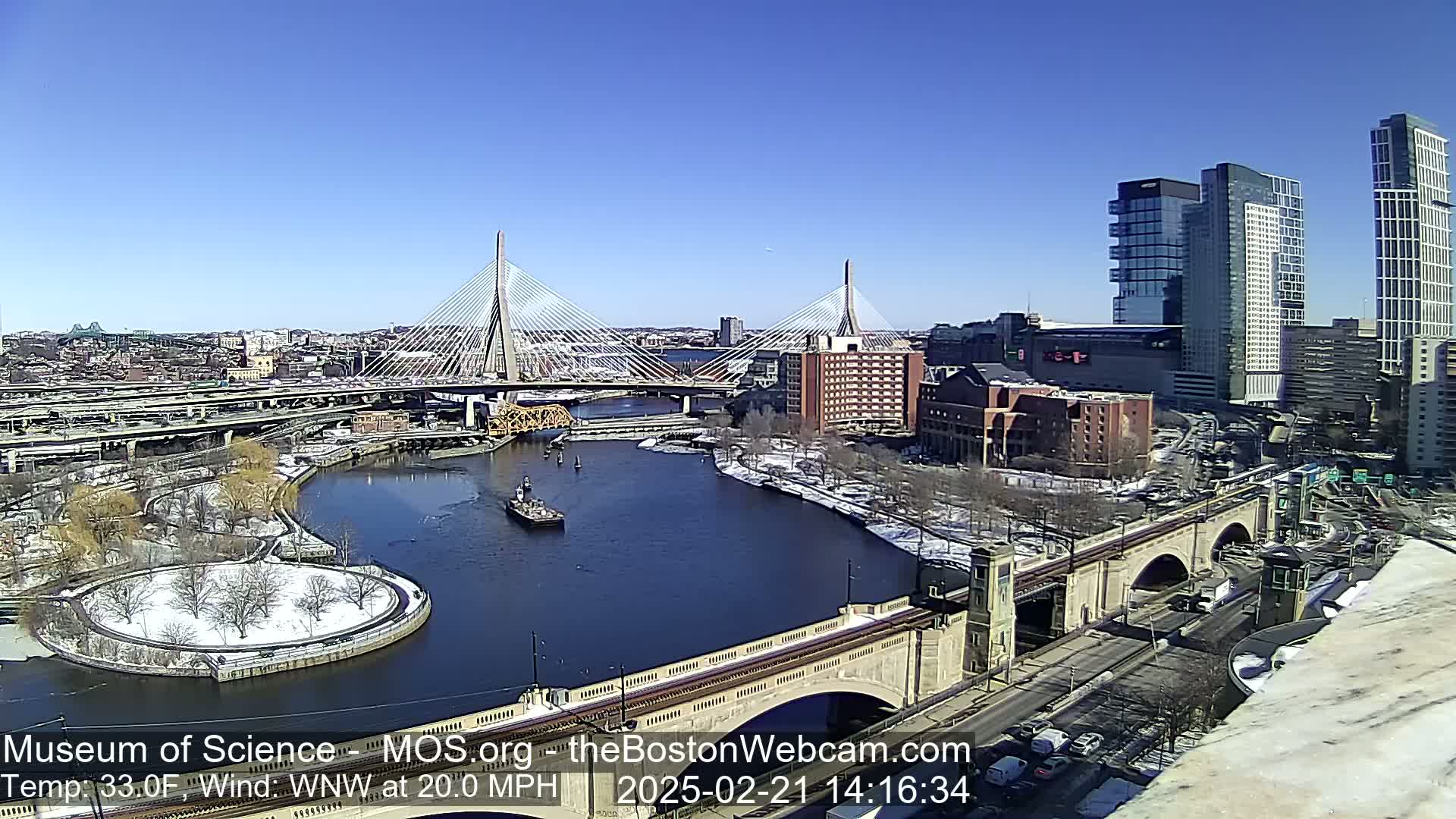 A sunny day with snow on the ground shows a cityscape with two cable-stayed bridges spanning a river, alongside modern and older buildings, under a clear blue sky.