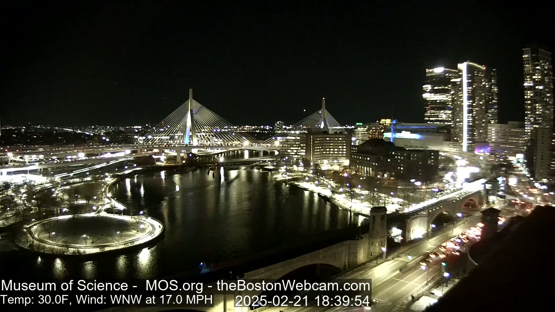 A nighttime aerial view of a city illuminated with lights, featuring two cable-stayed bridges spanning a river, with a wind of 17 mph from the west-northwest and a temperature of 30 degrees Fahrenheit.