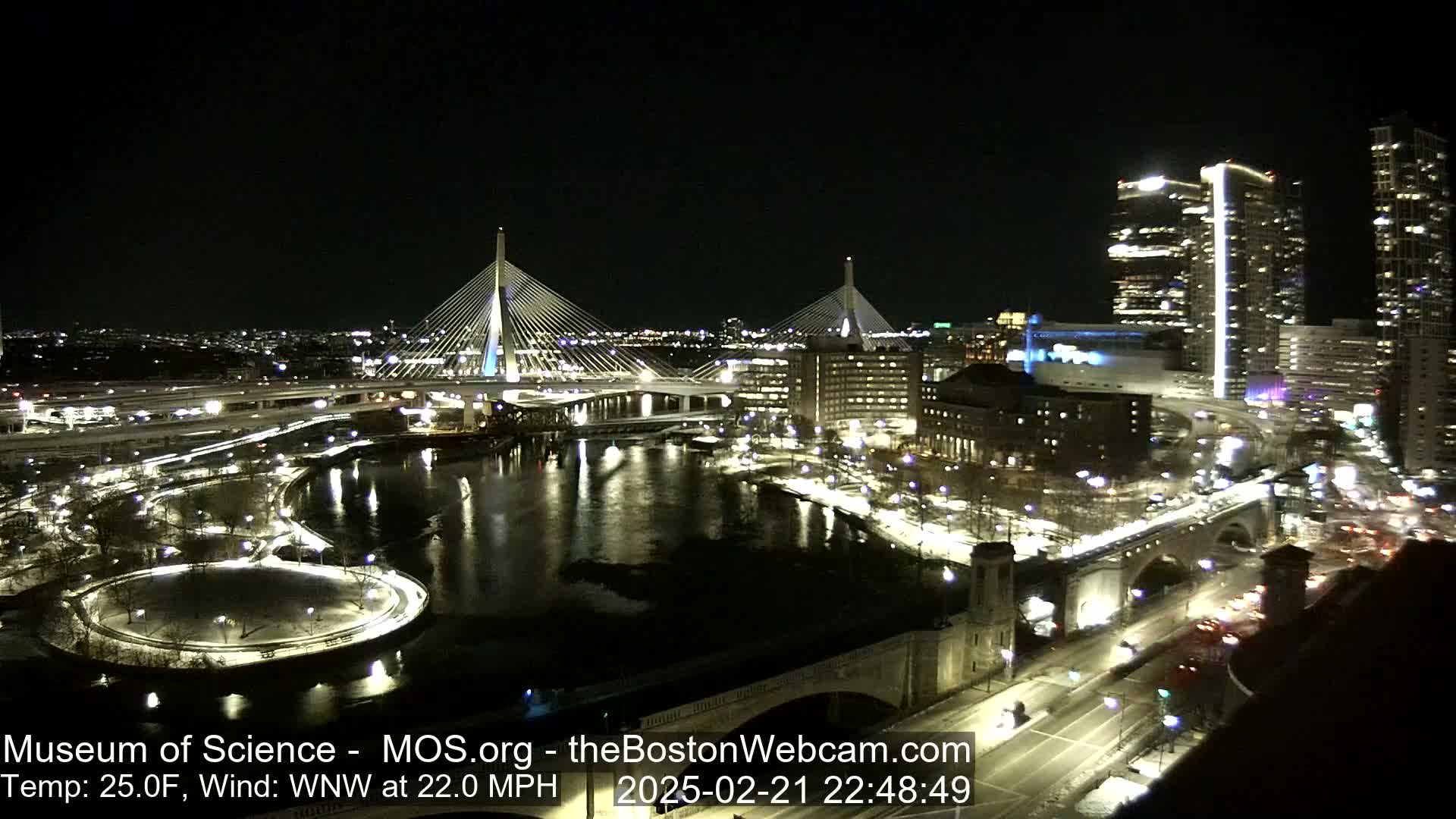 A nighttime view of a city illuminated by lights, featuring two cable-stayed bridges spanning a river, with a cold wind blowing at 22 mph.