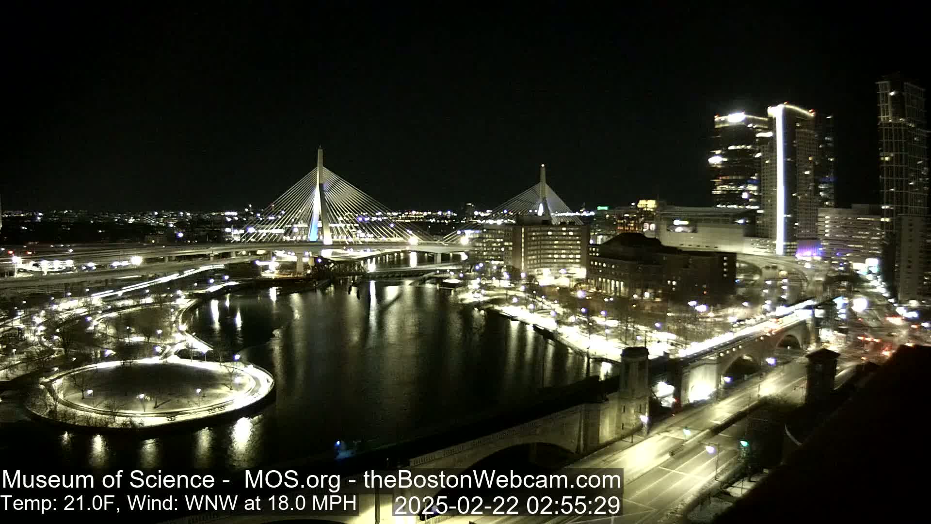 A nighttime view of two illuminated cable-stayed bridges spanning a dark river, with a city skyline and snow-covered ground under a clear sky, and a strong wind.